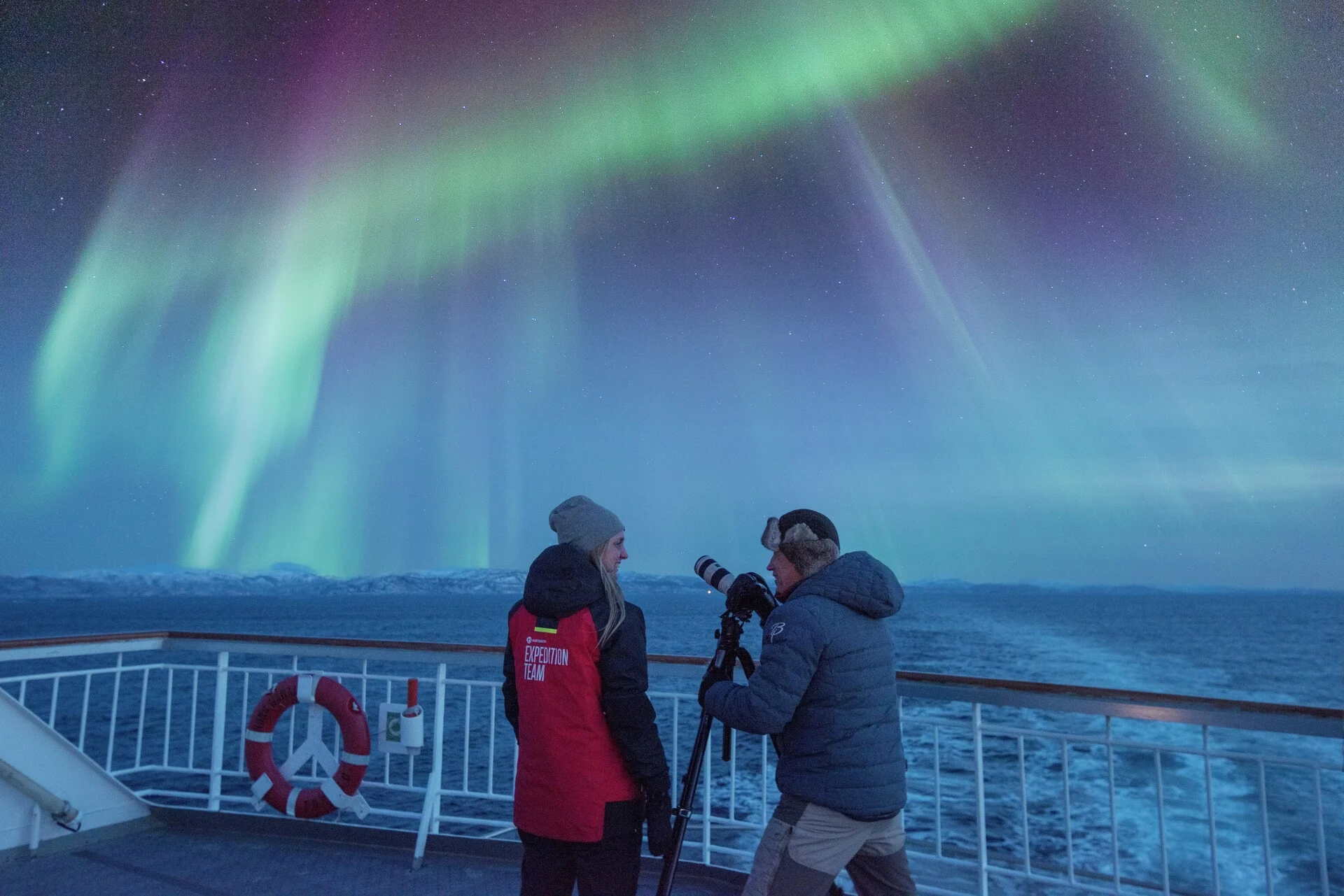 A tourist and crew member photographing the Northern Lights from the deck of a Hurtigruten ship