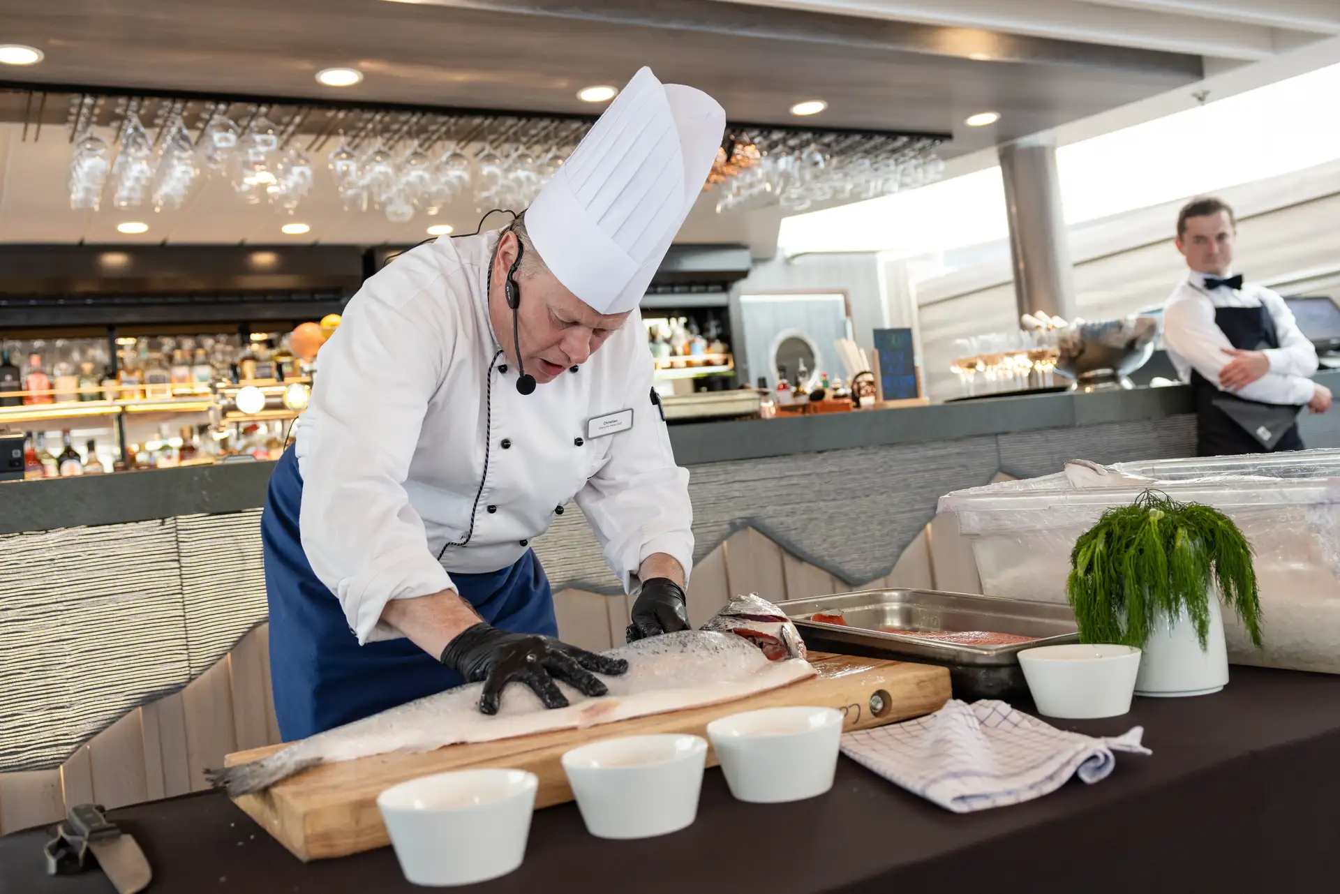 A chef wearing a white uniform and tall hat fillets a large fish on a cutting board in a modern kitchen, while a waiter watches nearby.