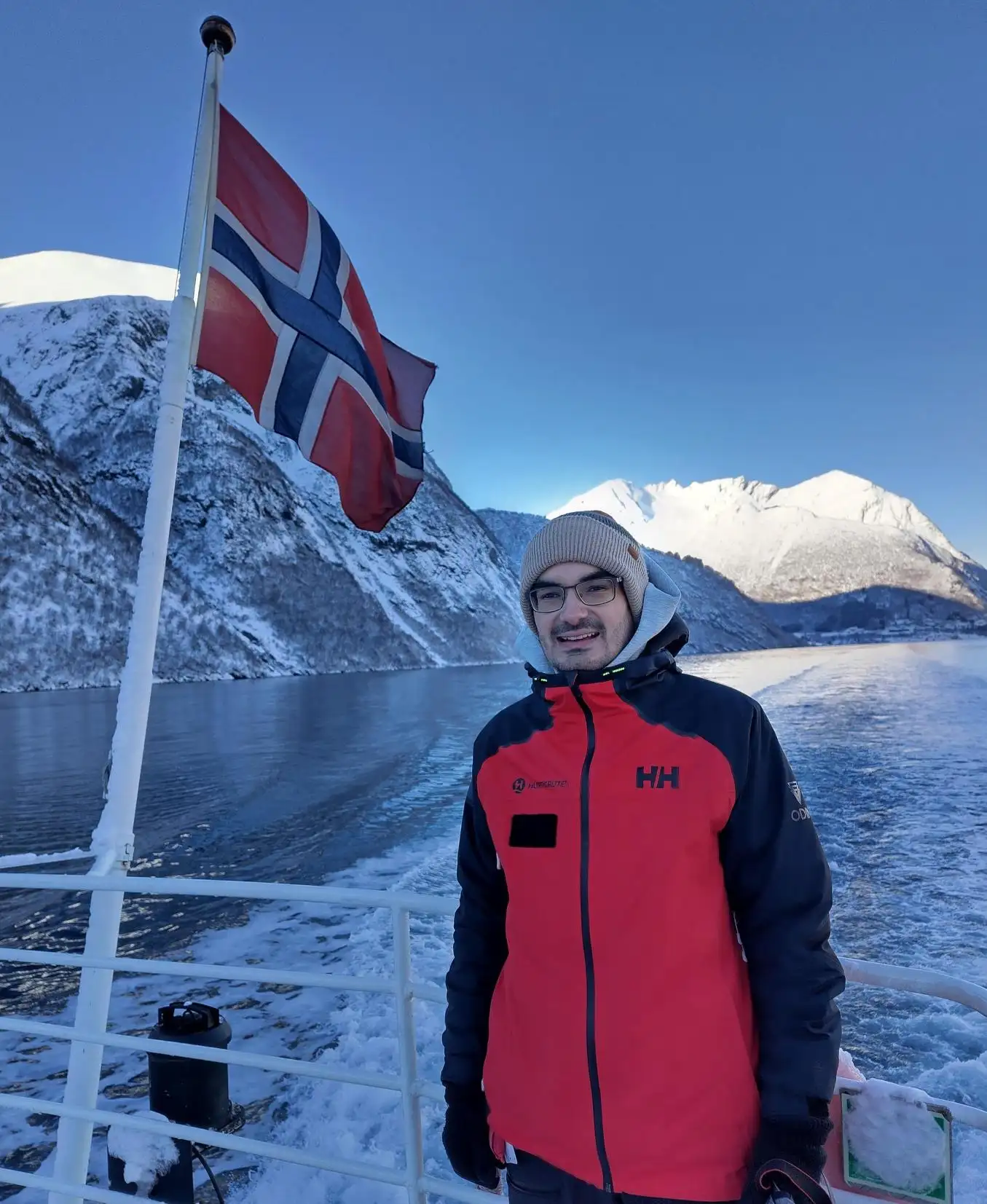 Expeditionsleiter Lukas Mohr steht im Winter an Deck eines Hurtigruten Schiffes. Neben ihm sieht man die norwegische Flagge.