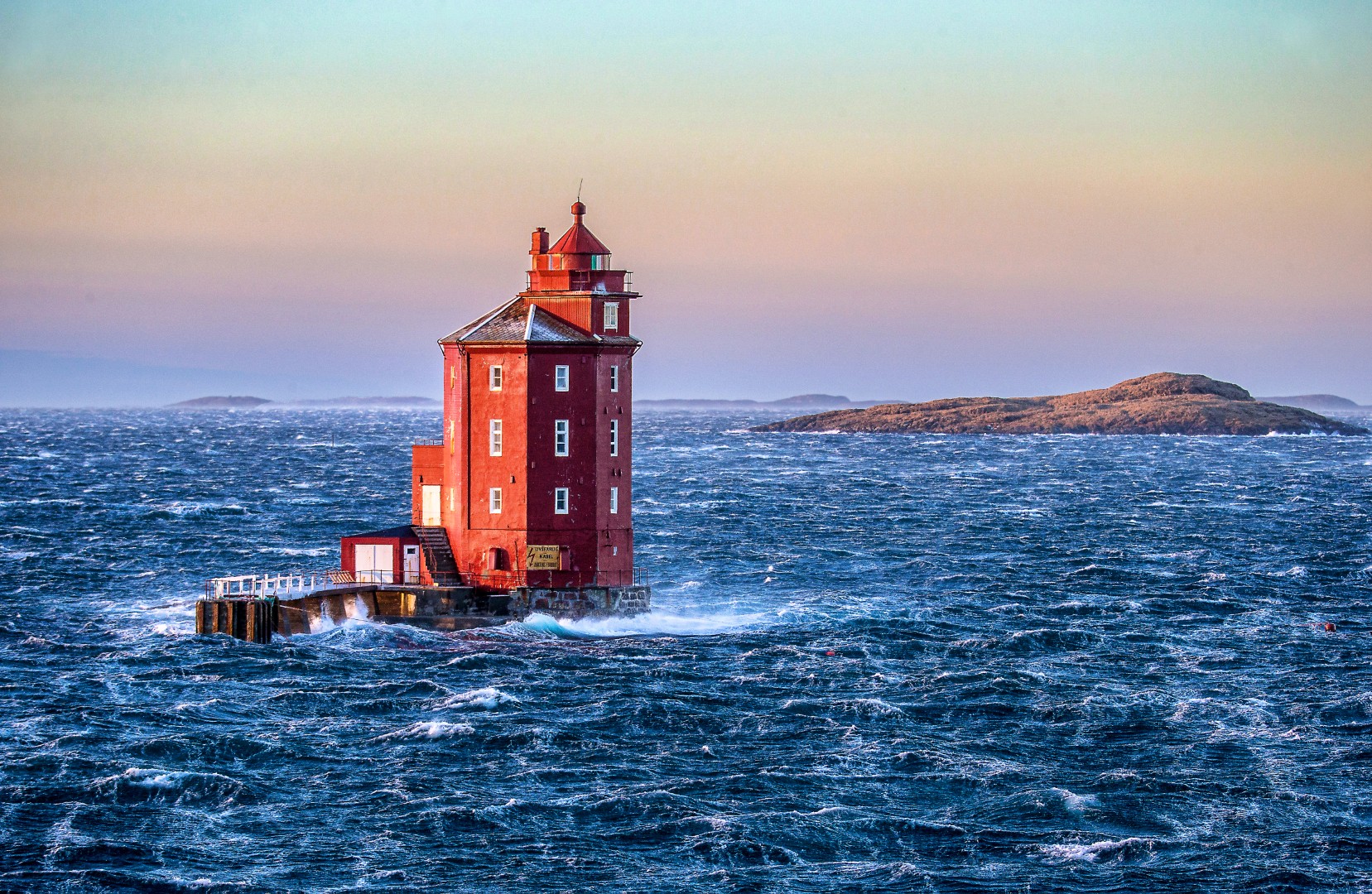 Ein roter Leuchtturm steht inmitten rauer, aufgewühlter See vor einem ruhigen Abendhimmel. In der Ferne sind Inseln zu sehen, die einen Kontrast zwischen Ruhe und Sturm bilden.