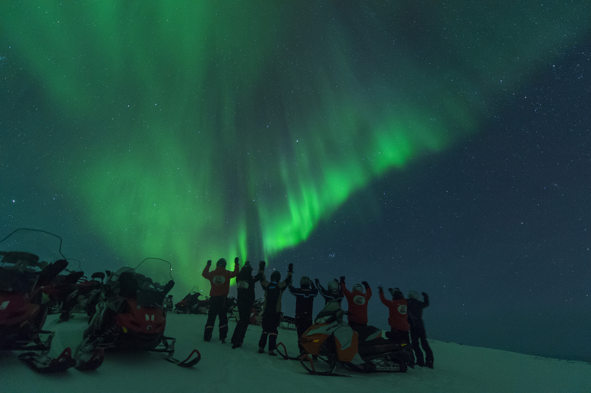 A group of people in winter gear stand on snow beside snowmobiles, arms raised, under a vibrant green aurora borealis in a starry night sky.