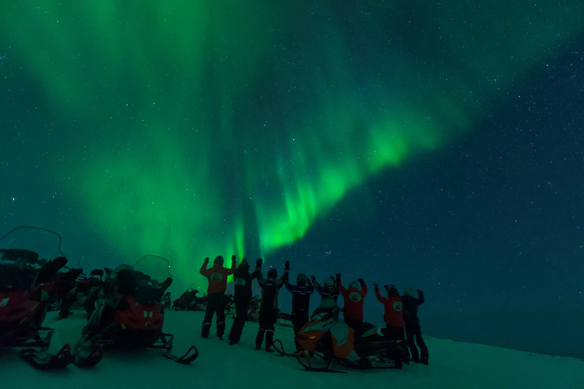 A group of people in winter gear stand on snow beside snowmobiles, arms raised, under a vibrant green aurora borealis in a starry night sky.