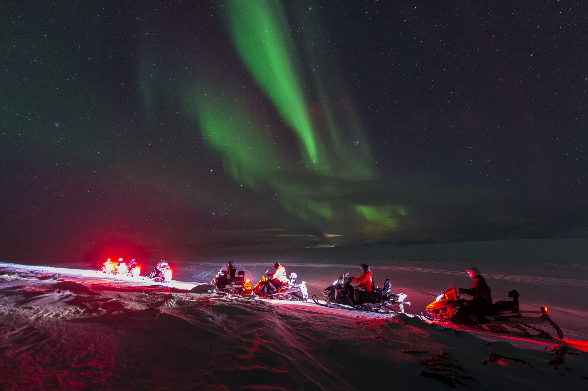 Snowmobilers in the foreground admire the vivid green Northern Lights in the starry night sky. A serene, awe-inspiring winter landscape.