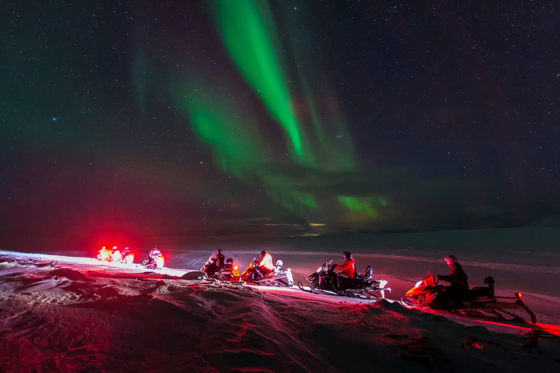 Snowmobilers in the foreground admire the vivid green Northern Lights in the starry night sky. A serene, awe-inspiring winter landscape.