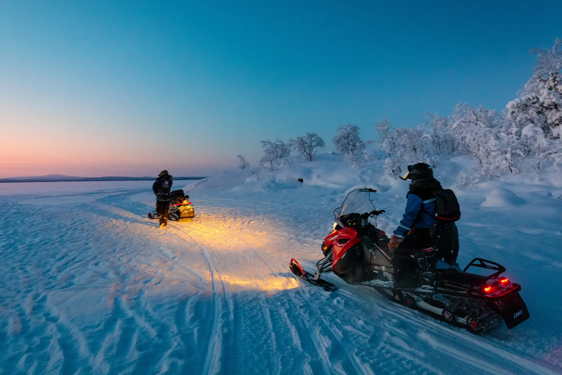 Two people on snowmobiles ride on a snow-covered landscape at dusk, with one leading and headlights illuminating the path. The sky is a gradient of blue and orange, creating a serene winter scene.