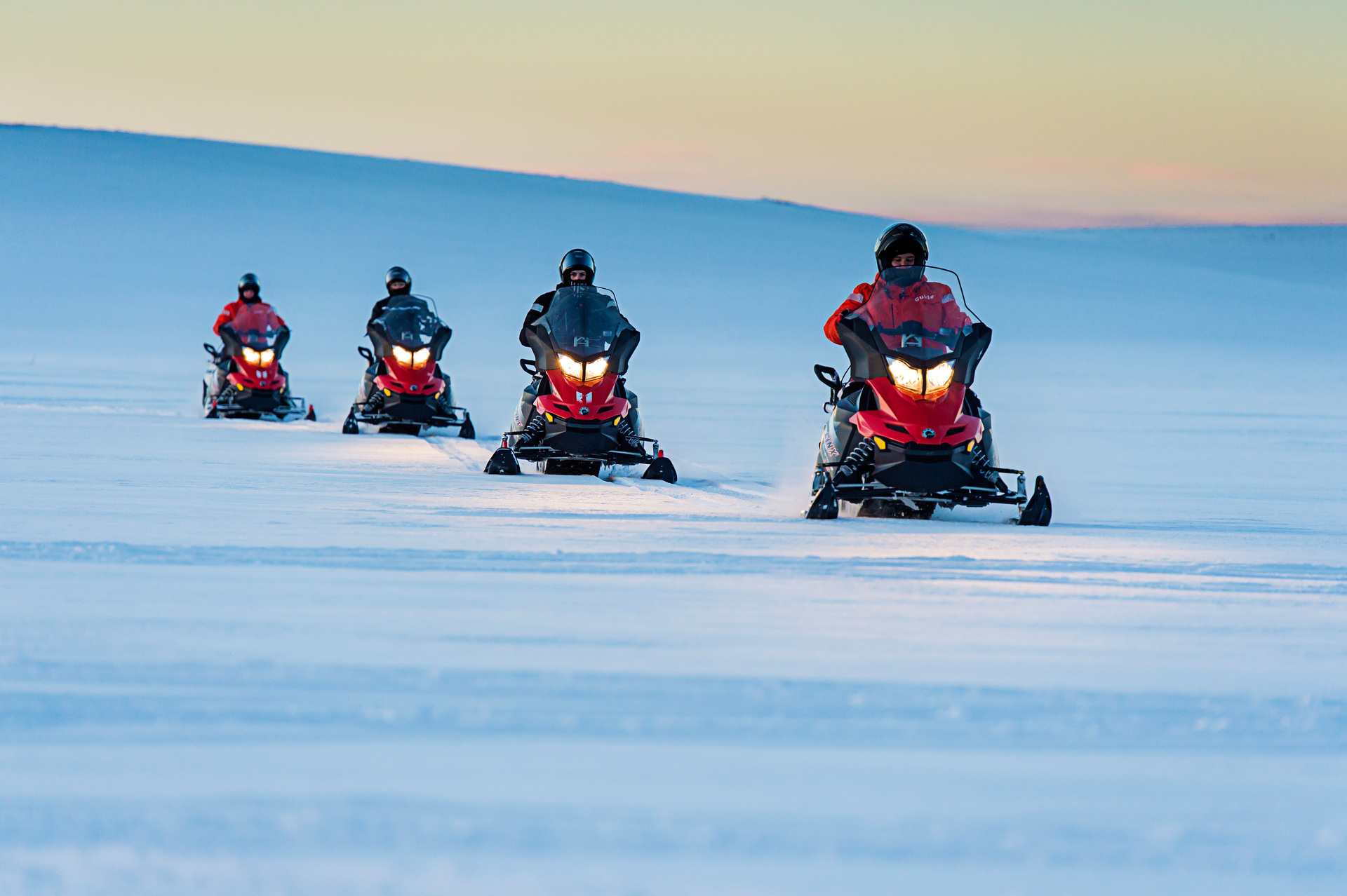 Four people riding red snowmobiles in a line across a vast snowy landscape under a clear sky during sunset. The scene conveys adventure and freedom.