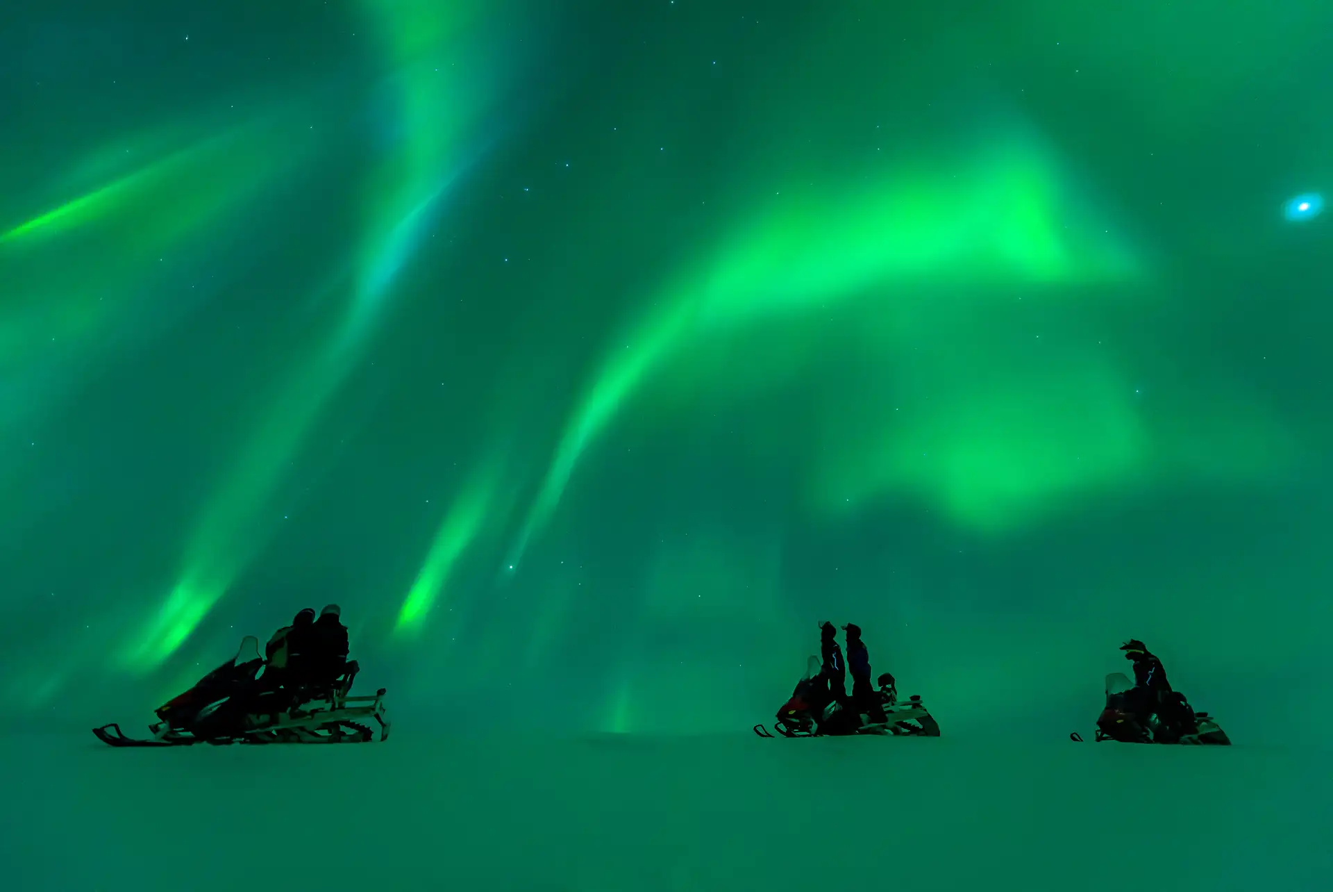 Snowmobilers watch the vibrant green Northern Lights in the night sky. The atmosphere is serene and awe-inspiring, with stars scattered above.