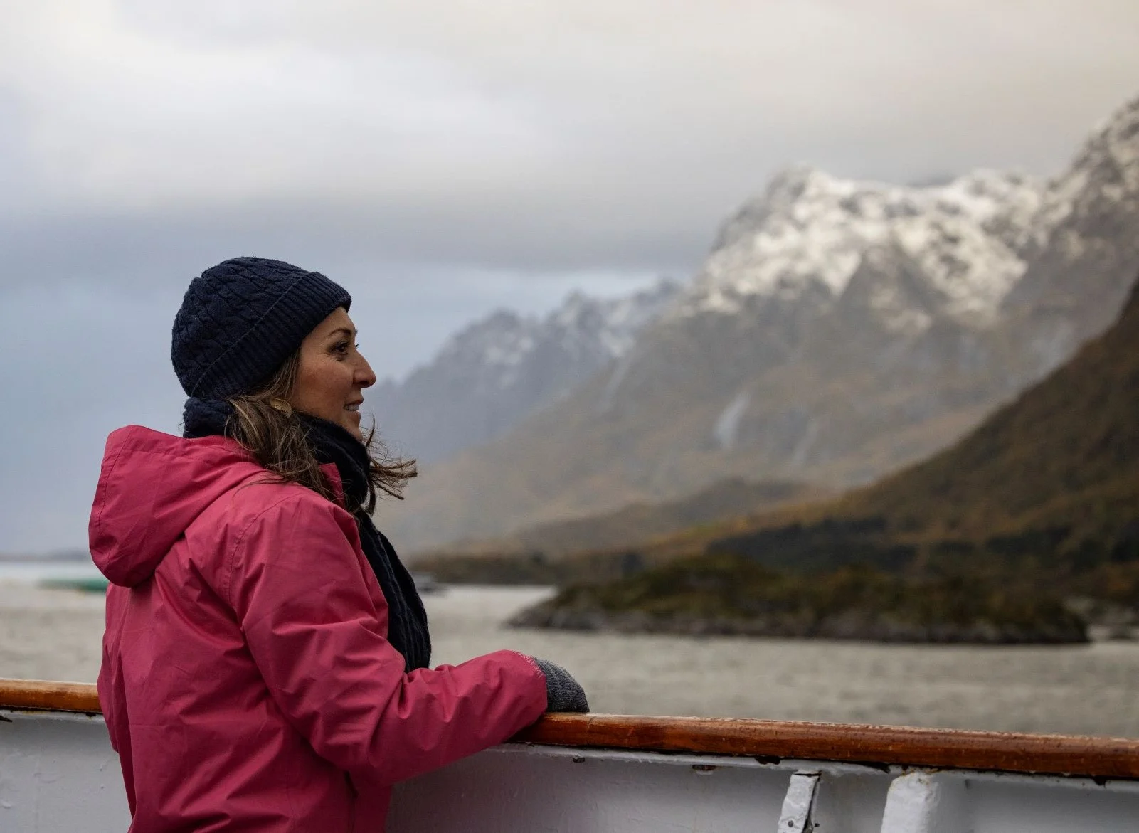 A woman in a pink jacket and navy beanie gazes at snow-capped mountains and a fjord while standing on a ship's deck.