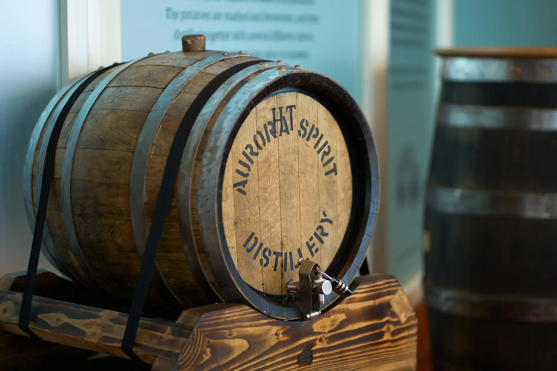 Ein kleines Eichenfass mit der Aufschrift „Aurora Spirit Distillery“ steht auf einer Holzhalterung mit einem Metallhahn und ist an Bord der MS Trollfjord von Hurtigruten ausgestellt. Im Hintergrund ist ein zweites Fass teilweise zu sehen, und an der Wand dahinter ist eine Informationstafel mit teilweise lesbarem Text über den Produktionsprozess der Brennerei angebracht – was darauf hindeutet, dass es sich hierbei um eine spezielle Spirituosen- oder Verkostungsstation an Bord handelt.