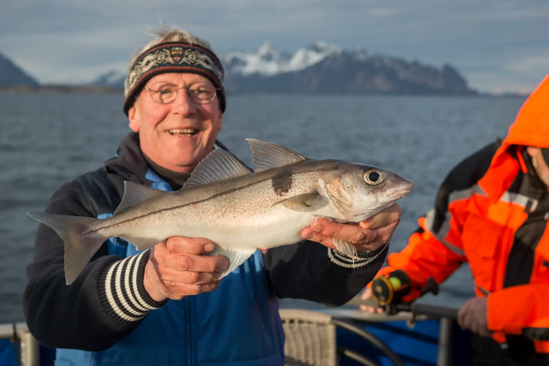 Smiling man in a blue jacket holds a large fish on a boat, with snowy mountains in the background. Another person in an orange jacket stands beside him.