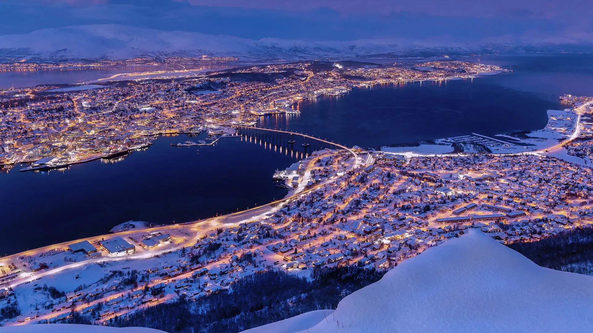 Snowy Tromsø cityscape at dusk, featuring warm lights reflecting off the water, surrounded by snow-covered mountains and a serene, twilight sky.