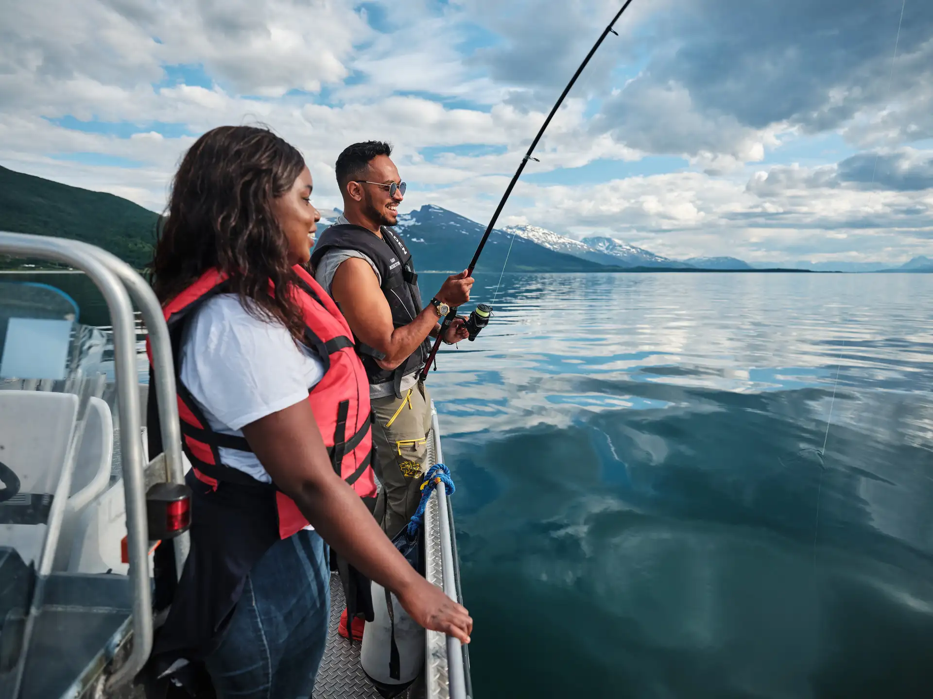 A man and woman in life jackets fish from a boat on a calm lake surrounded by mountains. The mood is peaceful and focused under a partly cloudy sky.