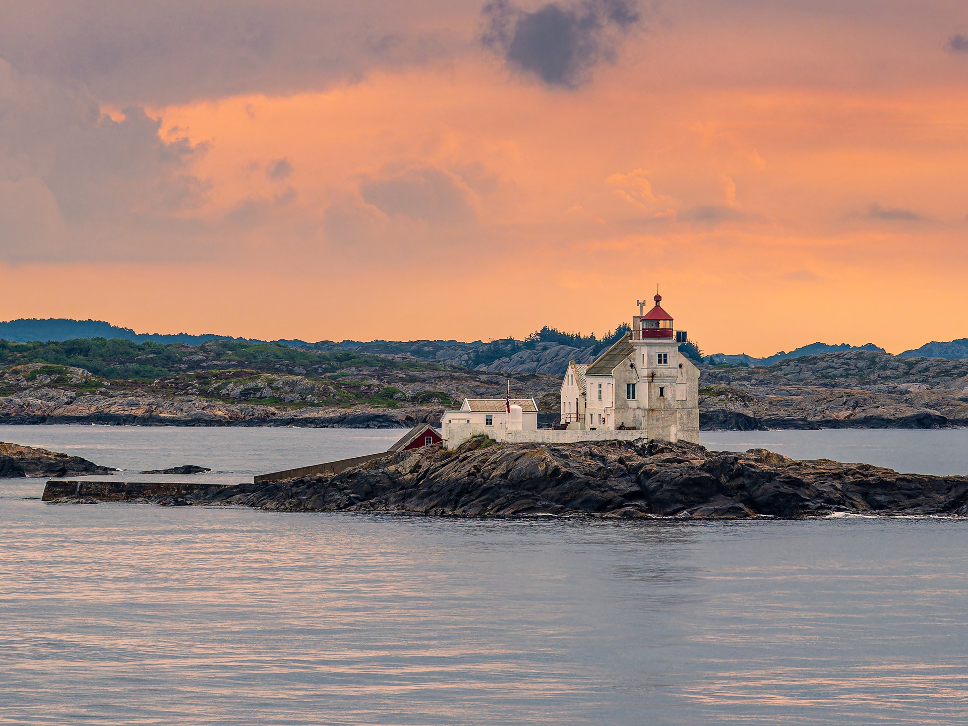 A solitary, white lighthouse sits on a rocky islet, surrounded by calm waters. The sky glows with a warm, orange sunset, creating a serene ambiance.