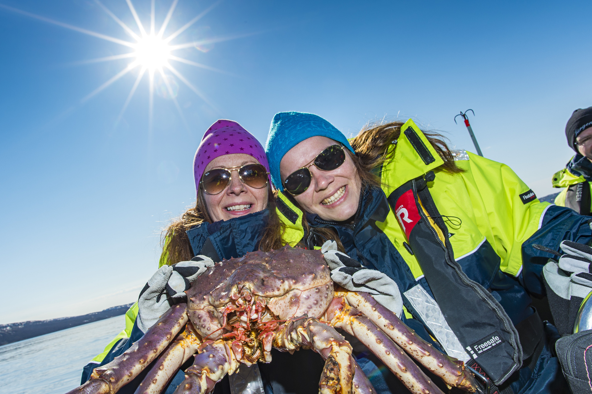 Two people in bright winter jackets and sunglasses smile broadly while holding a large crab under a clear blue sky with the sun shining brightly.