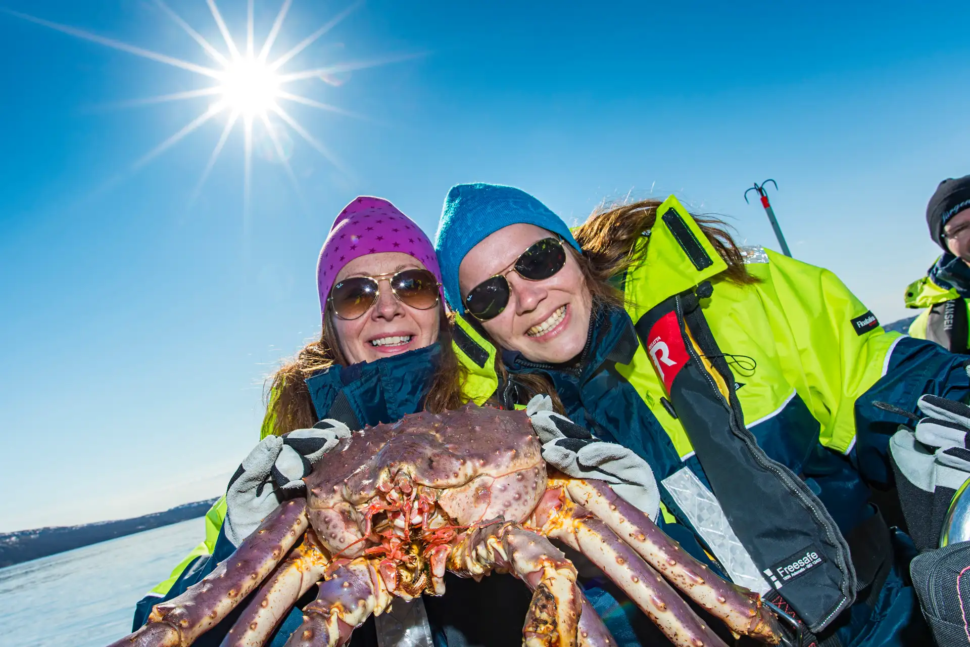 Two people in bright winter jackets and sunglasses smile broadly while holding a large crab under a clear blue sky with the sun shining brightly.