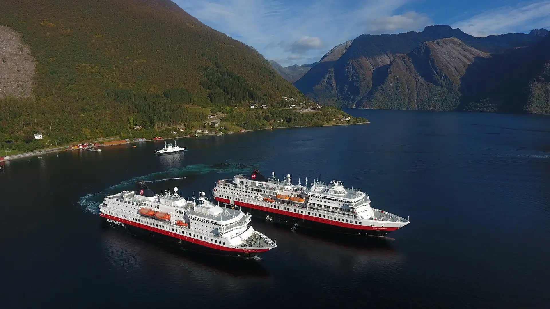 Aerial view of Hurtigruten's MS Nordkapp and MS Finnmarken passing each other in the Horundfjord, with autumn-forested mountains and a small coastal settlement visible behind