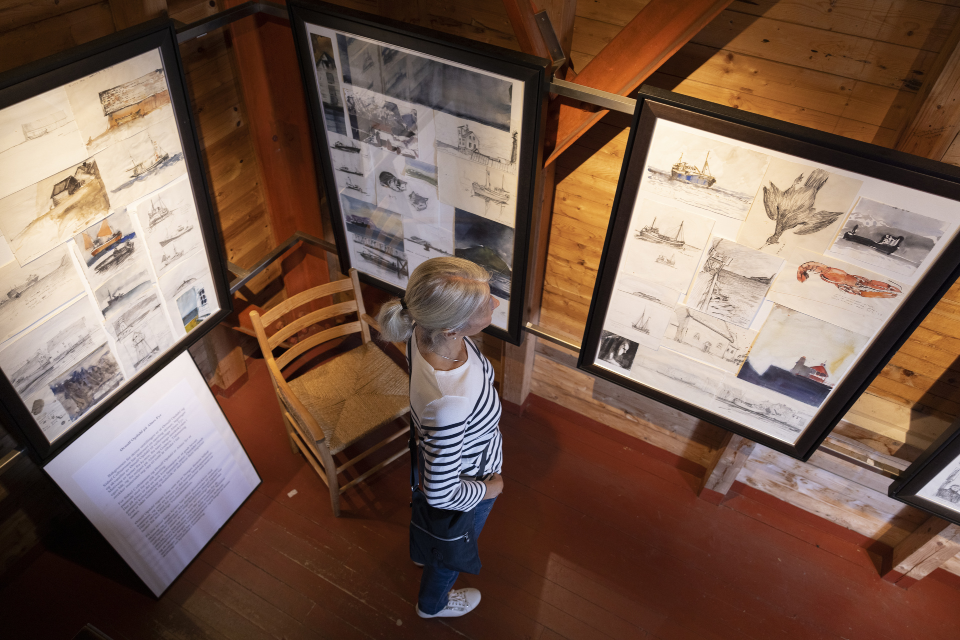 A woman with blonde hair views maritime-themed sketches in a wooden gallery. The artworks are framed, and the setting feels cozy and contemplative.