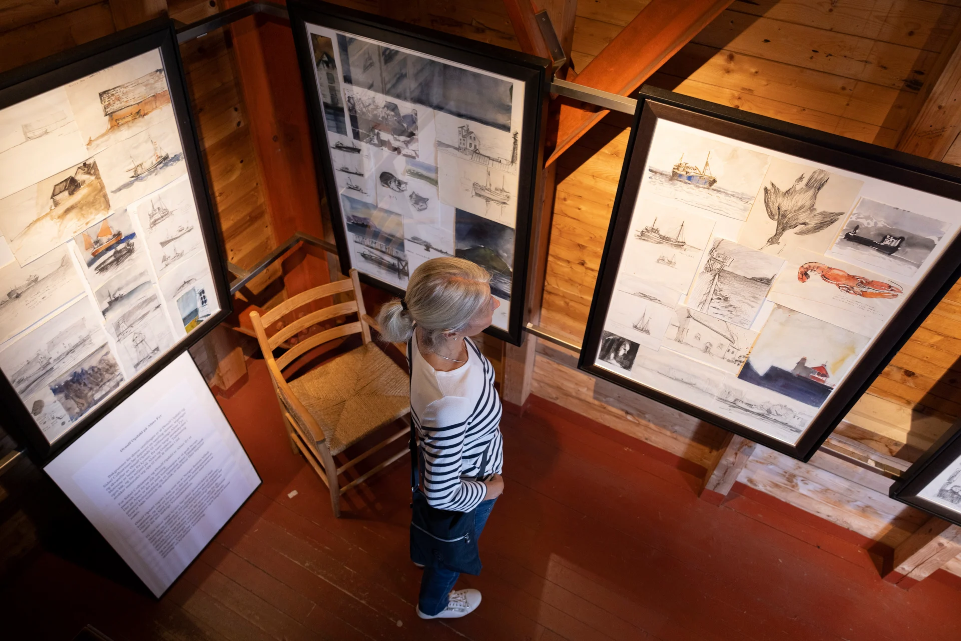 A woman with blonde hair views maritime-themed sketches in a wooden gallery. The artworks are framed, and the setting feels cozy and contemplative.