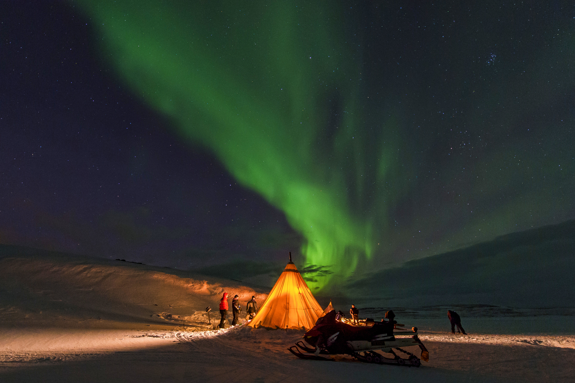 A glowing orange tent in snowy terrain under a vibrant green aurora borealis. People stand outside, creating a serene, awe-inspired scene. Stars dot the clear night sky.