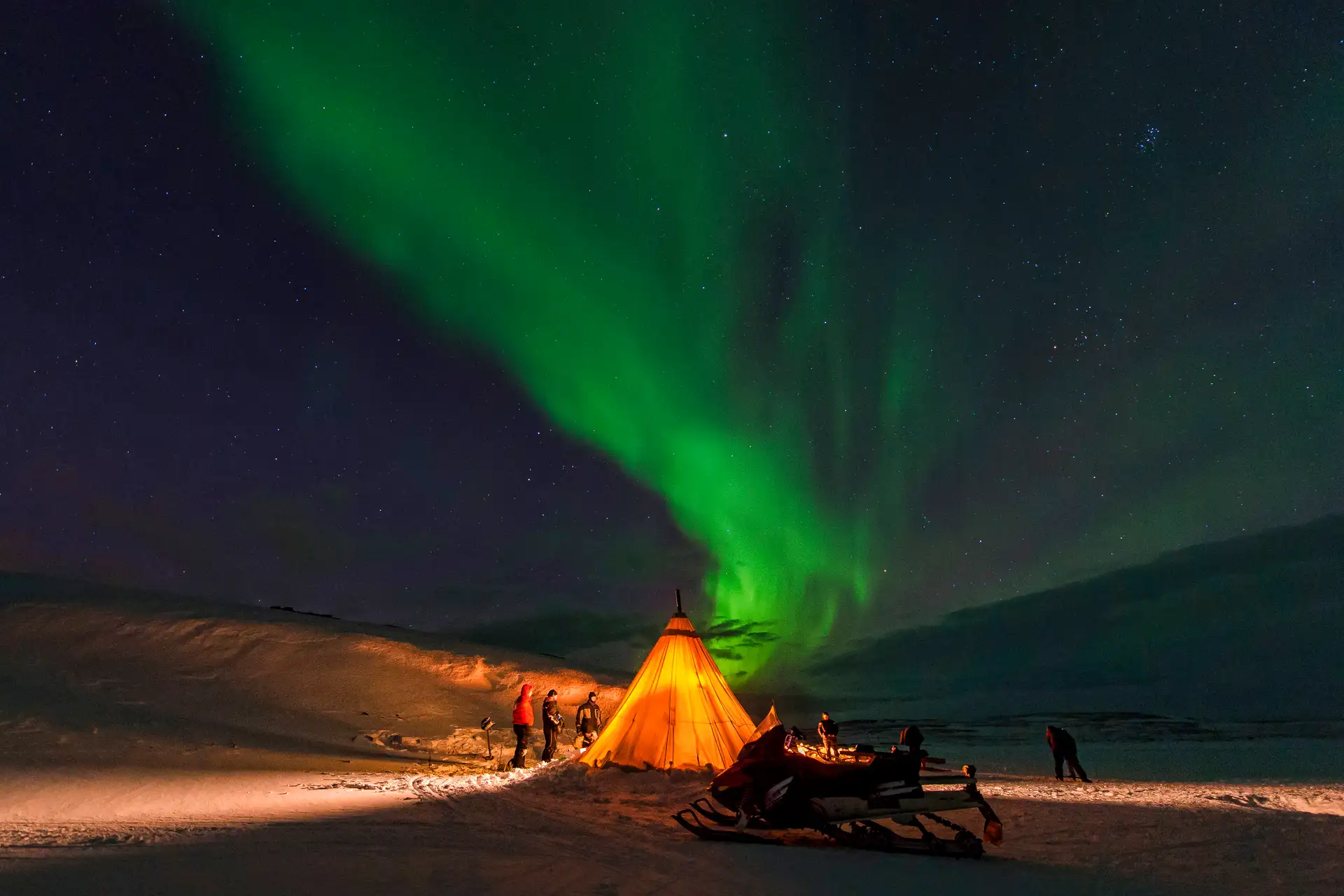 A glowing orange tent in snowy terrain under a vibrant green aurora borealis. People stand outside, creating a serene, awe-inspired scene. Stars dot the clear night sky.