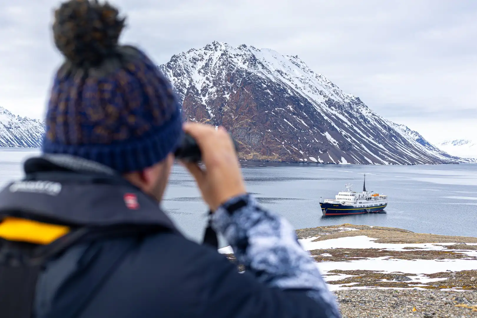 Ein Gast blickt durch ein Fernglas auf das Expeditionsschiff MS Serenissima von Hurtigruten Svalbard, das in einem Fjord auf Spitzbergen vor Anker liegt. Der Blick erfolgt von einem leicht verschneiten Ufer aus, während sich im Hintergrund ein dramatischer, von Schneestreifen durchzogener Berg erhebt.