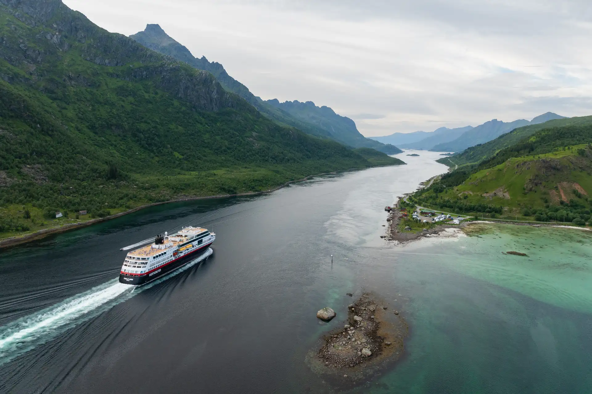 Luftaufnahme eines Hurtigruten Schiffes, das durch einen schmalen Fjord fährt, umgeben von üppig grünen Bergen und ruhigem Wasser. Die Szene wirkt friedlich und majestätisch.