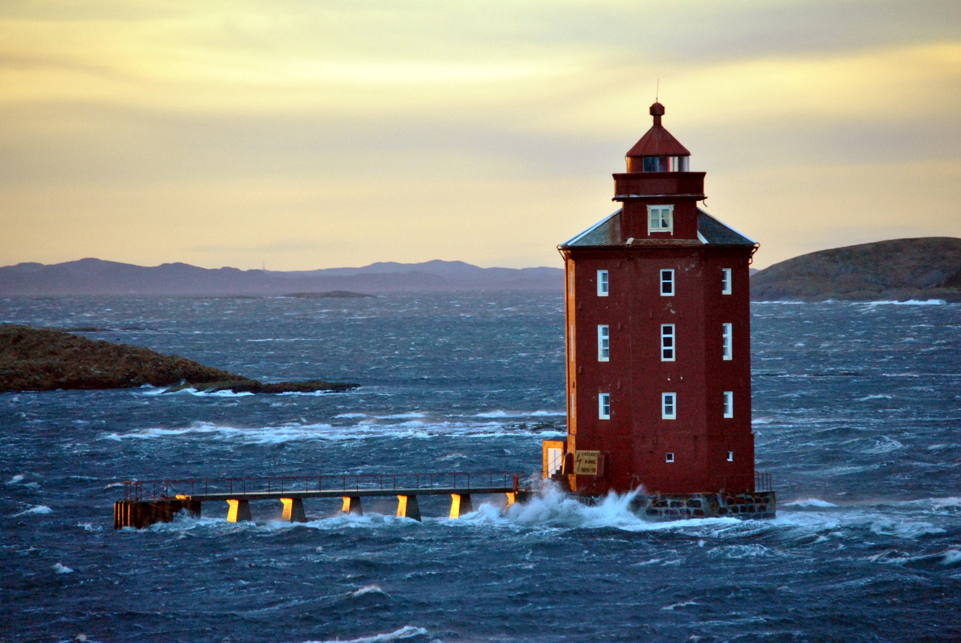 Ein roter Leuchtturm steht unerschütterlich inmitten tosender Wellen unter einem goldenen Himmel und vermittelt ein Gefühl von Standhaftigkeit und Einsamkeit. Im Hintergrund sind Hügel zu sehen.
