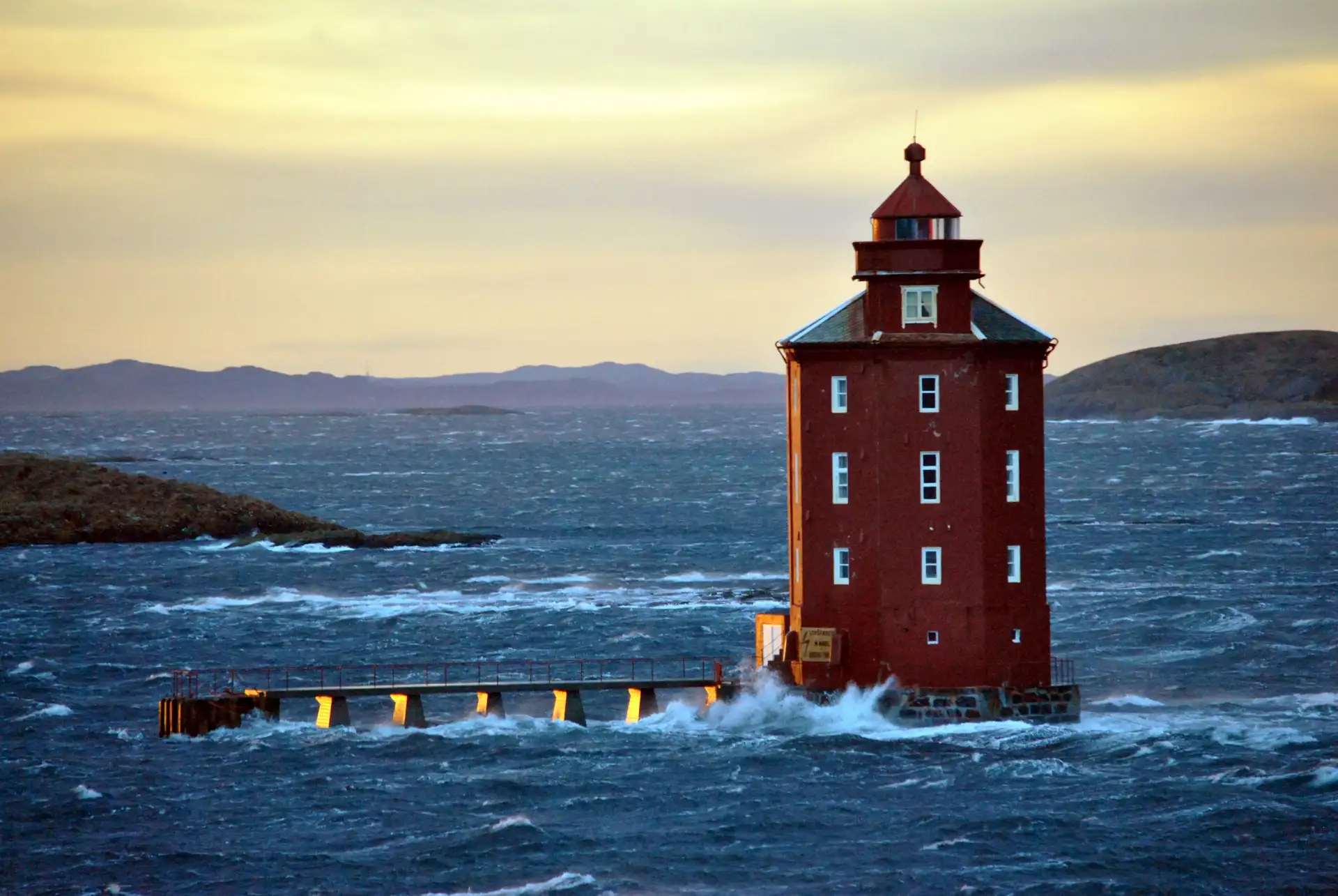 Ein roter Leuchtturm steht unerschütterlich inmitten tosender Wellen unter einem goldenen Himmel und vermittelt ein Gefühl von Standhaftigkeit und Einsamkeit. Im Hintergrund sind Hügel zu sehen.