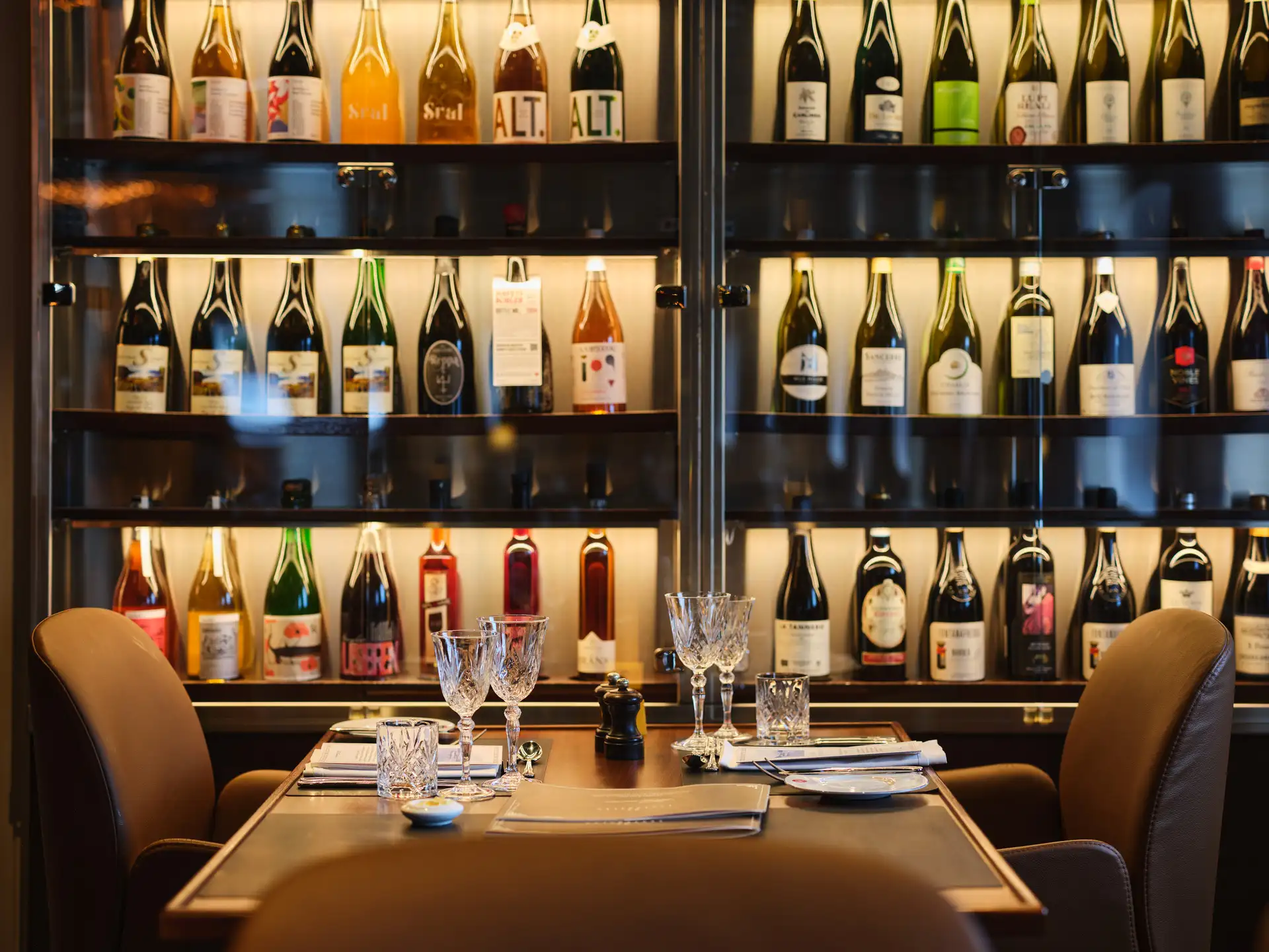 A set dining table with crystal glassware and menus in the Torget Restaurant aboard MS Vesterålen, with a backlit floor-to-ceiling wine display cabinet behind
