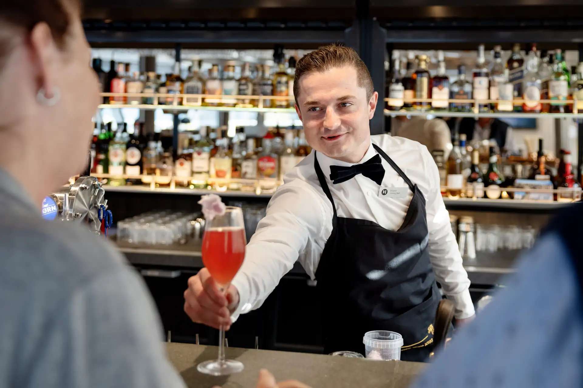 A bartender serving a pink cocktail in a champagne flute to a guest across the bar at the 1893 Bar aboard MS Finnmarken, with a well-stocked back bar behind