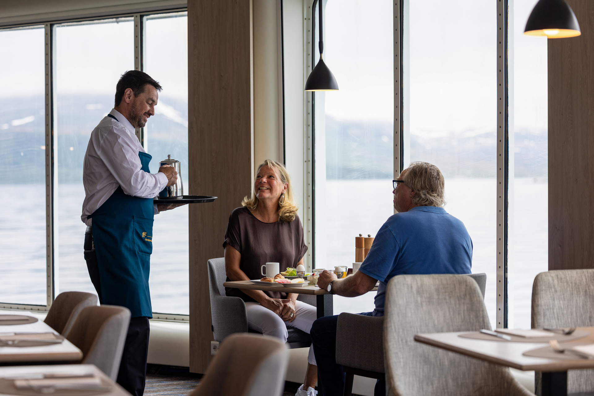 A waiter serves coffee to a smiling couple at a table in a cozy restaurant with large windows overlooking a scenic lake and mountains. Relaxed atmosphere.
