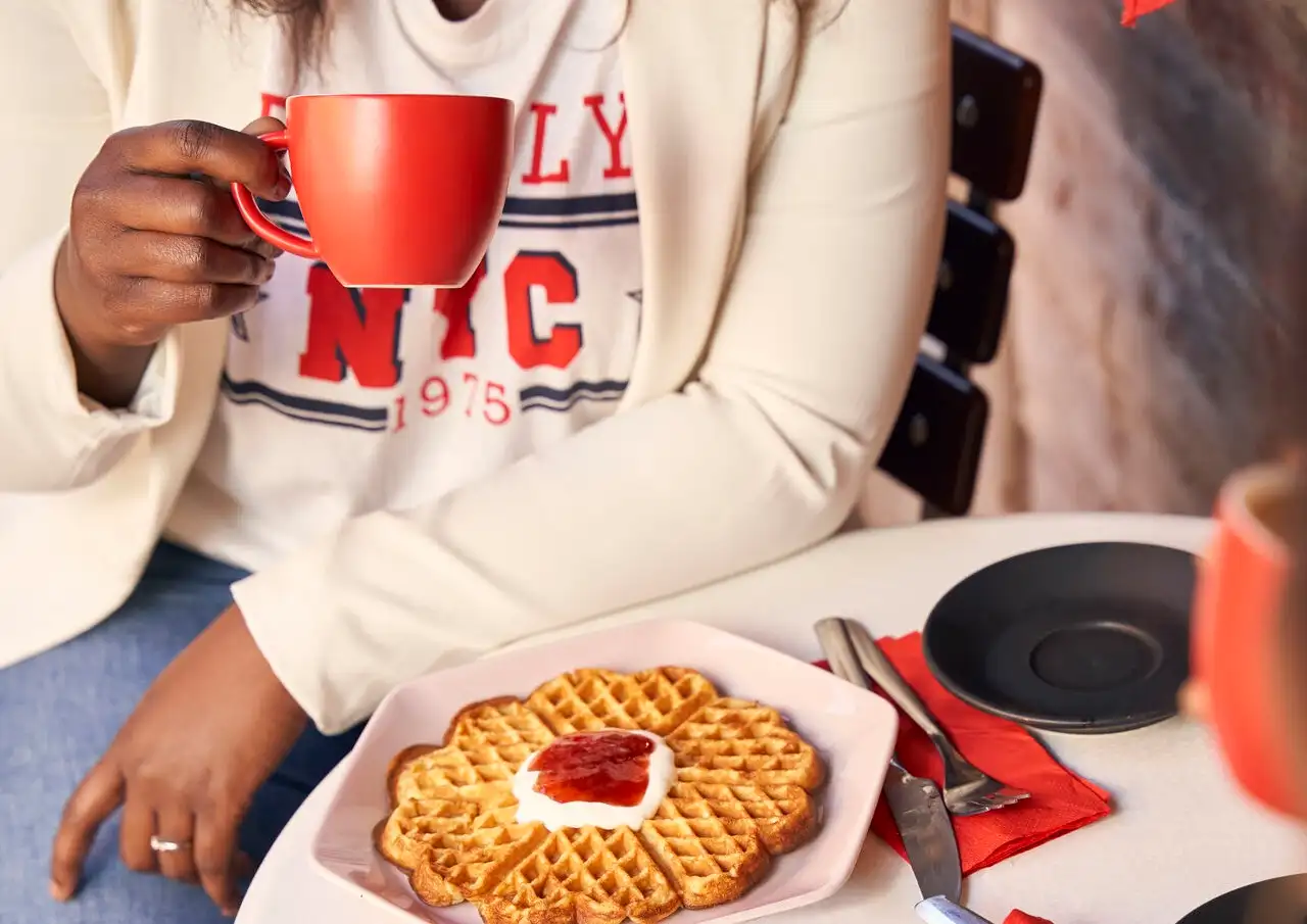 Ein Gast hält in einem Café in Bergen, Norwegen, eine rote Kaffeetasse mit herzförmigen norwegischen Waffeln auf einem rosa Teller in der Hand.
