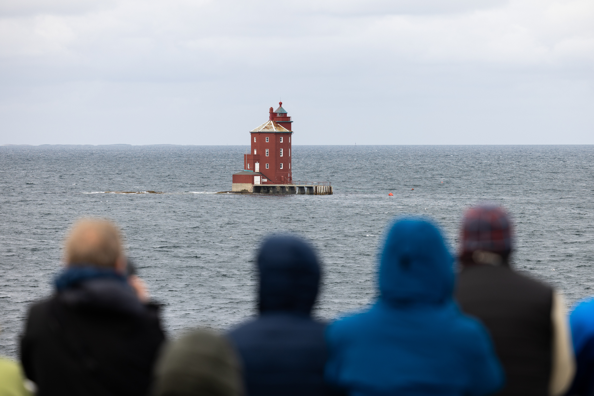 Menschen in Jacken stehen an Deck und betrachten einen einsamen roten Leuchtturm auf einem ruhigen, weiten Meer unter einem bewölkten Himmel, was ein Gefühl von Neugier und Gelassenheit vermittelt.