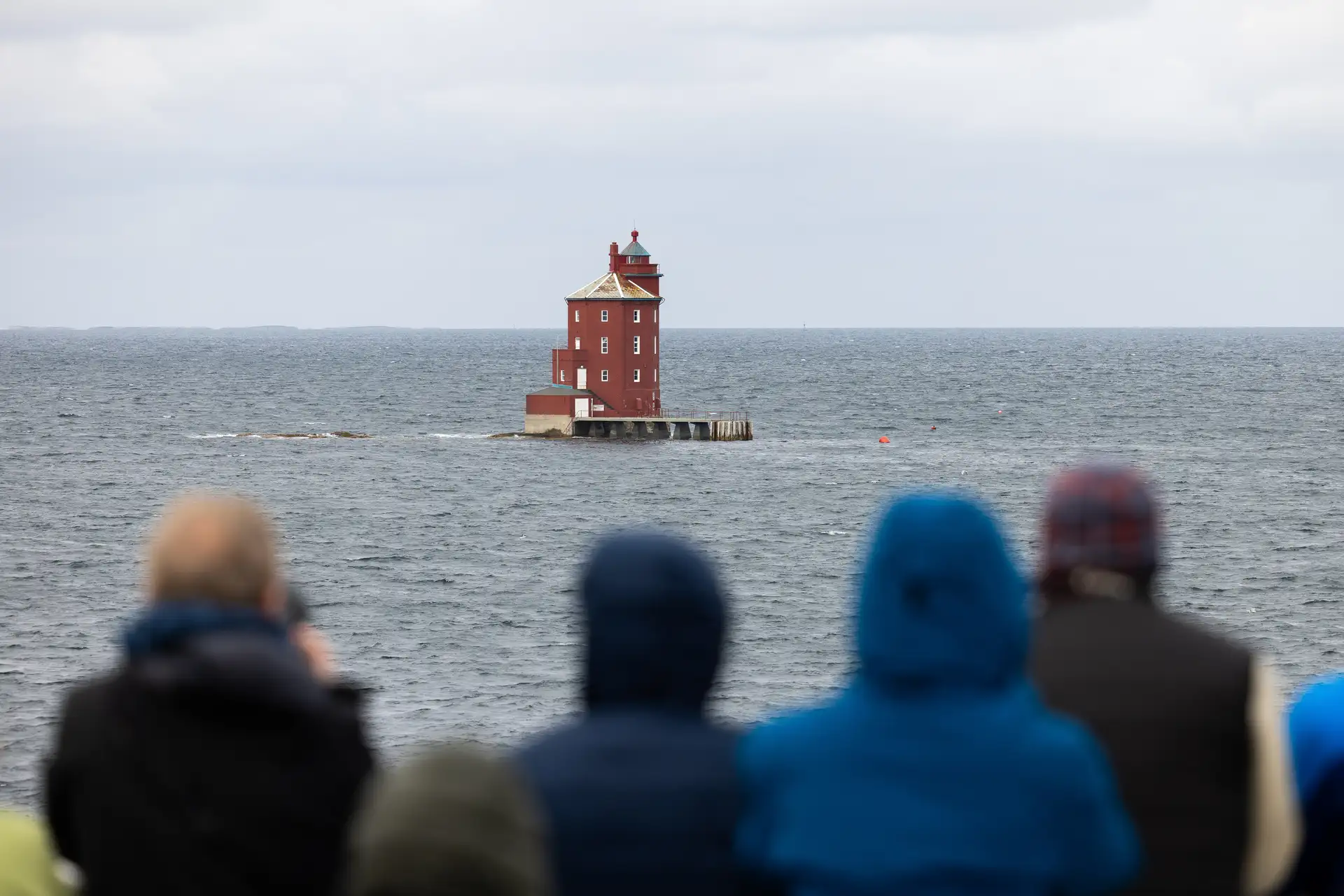 Menschen in Jacken stehen an Deck und betrachten einen einsamen roten Leuchtturm auf einem ruhigen, weiten Meer unter einem bewölkten Himmel, was ein Gefühl von Neugier und Gelassenheit vermittelt.