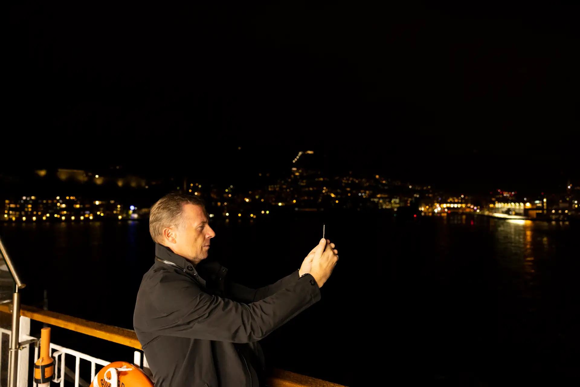 A guest photographing a Norwegian coastal town at night from the outdoor deck of Hurtigruten's MS Richard With, with harbour lights reflected in the dark water below