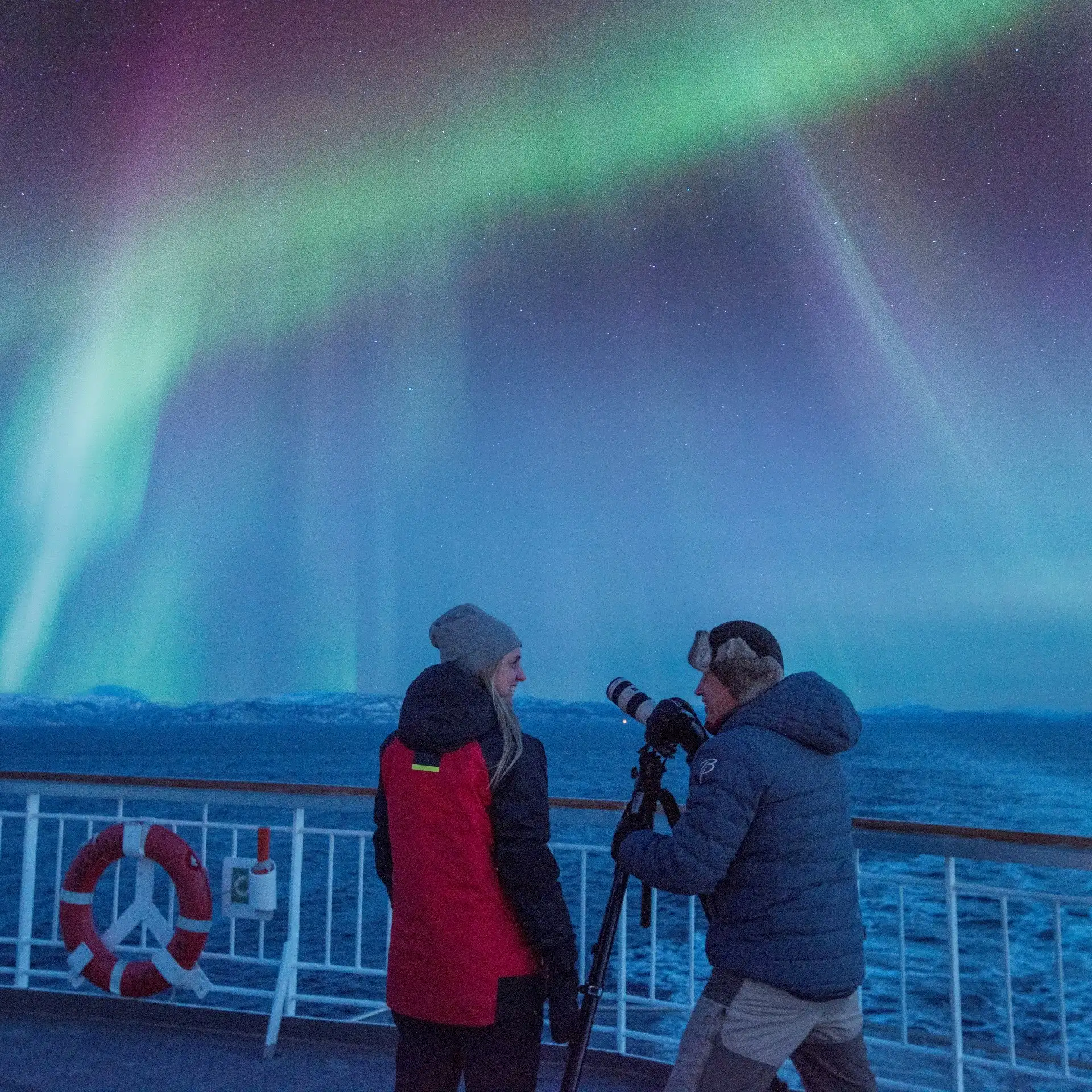 A boy and a girl is standing on Hurtigruten's ship looking at the sky - it is lit up in green, red  and blue