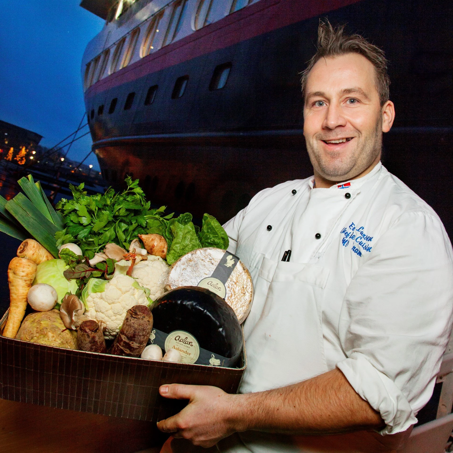A Hurtigruten Chef about to board the ship. He is holding a big tray of local goods. Vegetables and cheese mostly.