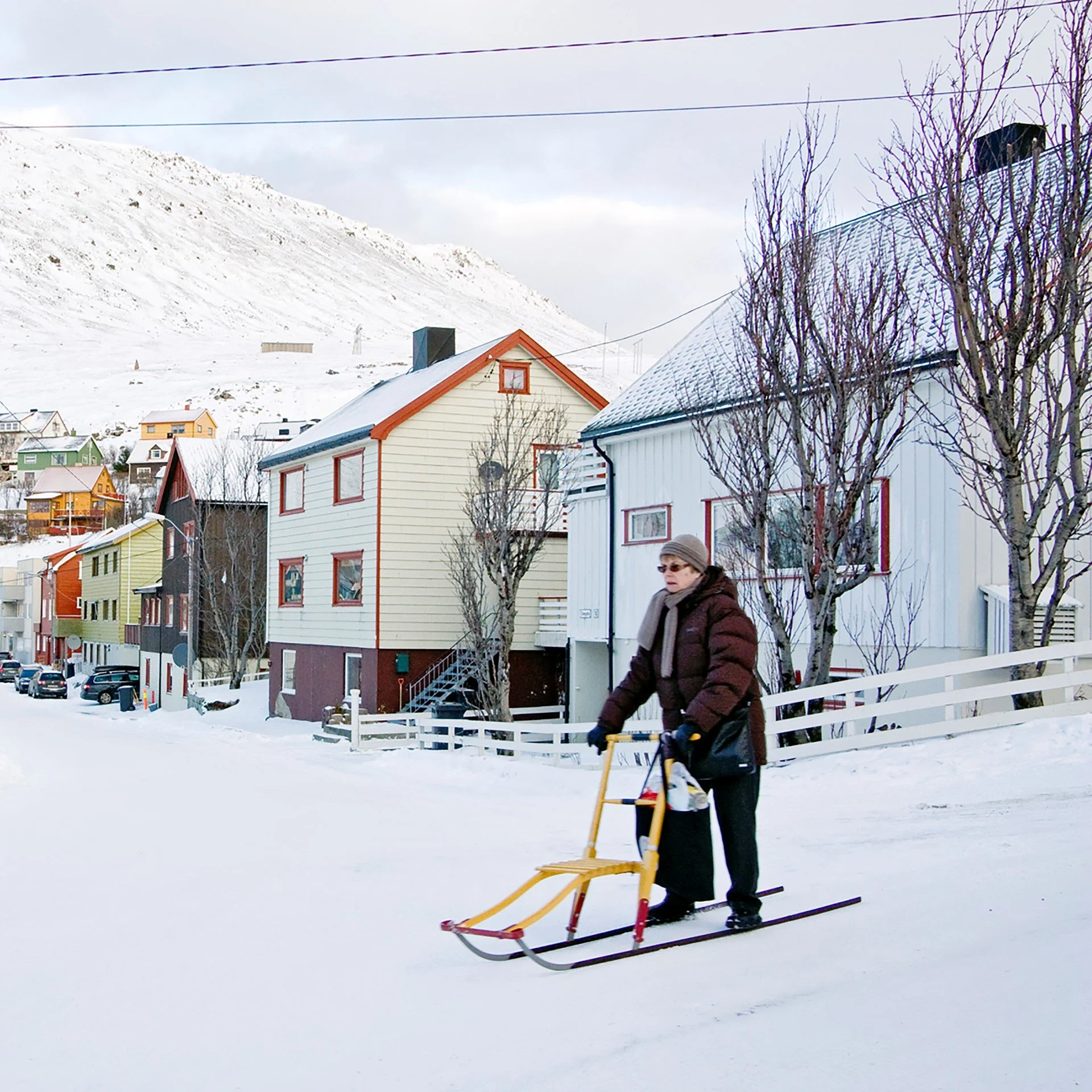 Photo showing an elderly woman in the snow, behind her a street with beautiful and colorful, small wooden houses