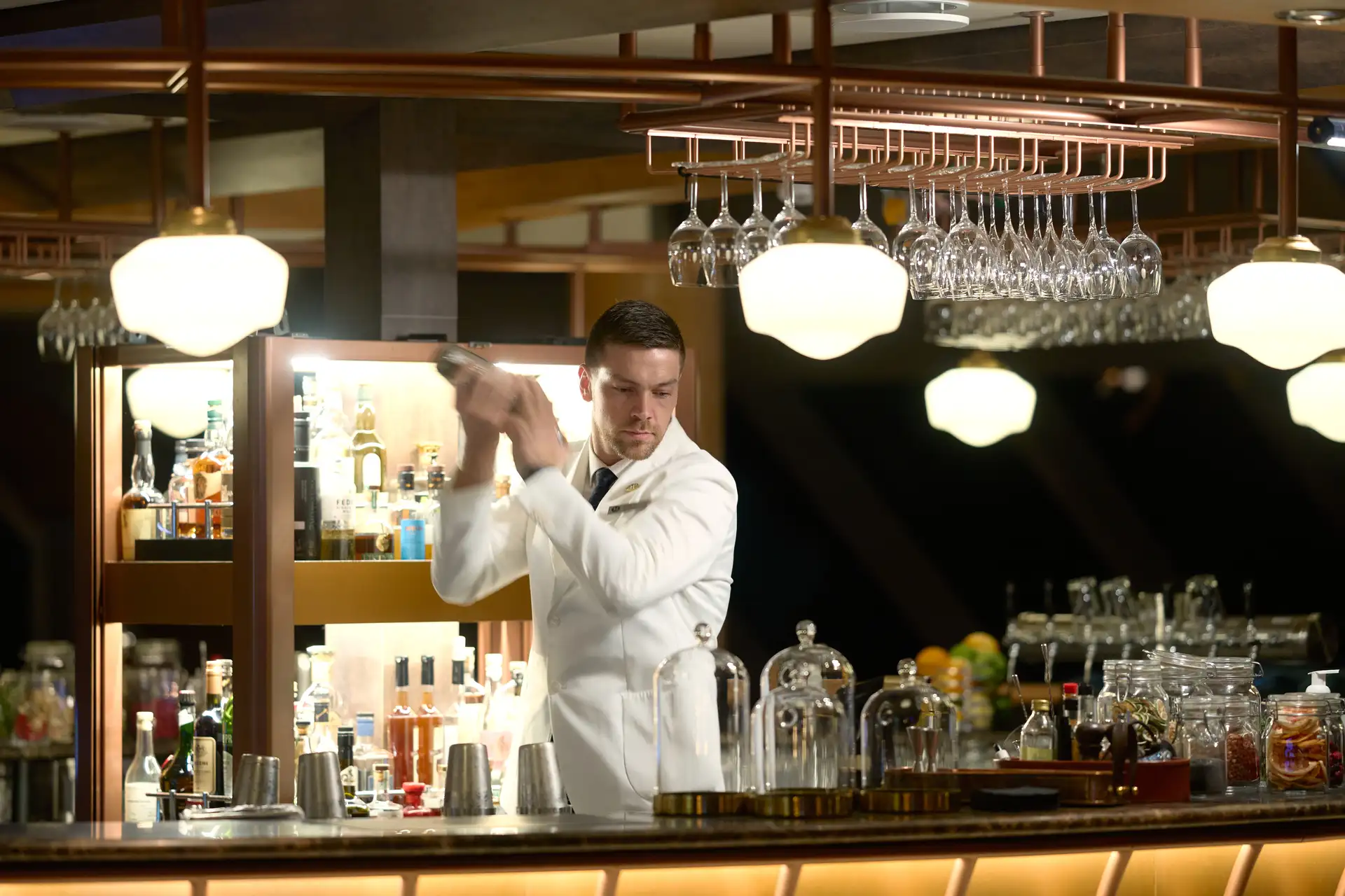 A bartender in a white jacket shaking a cocktail behind the bar of the 1893 Bar aboard a Hurtigruten ship, with rows of spirits, hanging glassware, and copper fixtures overhead