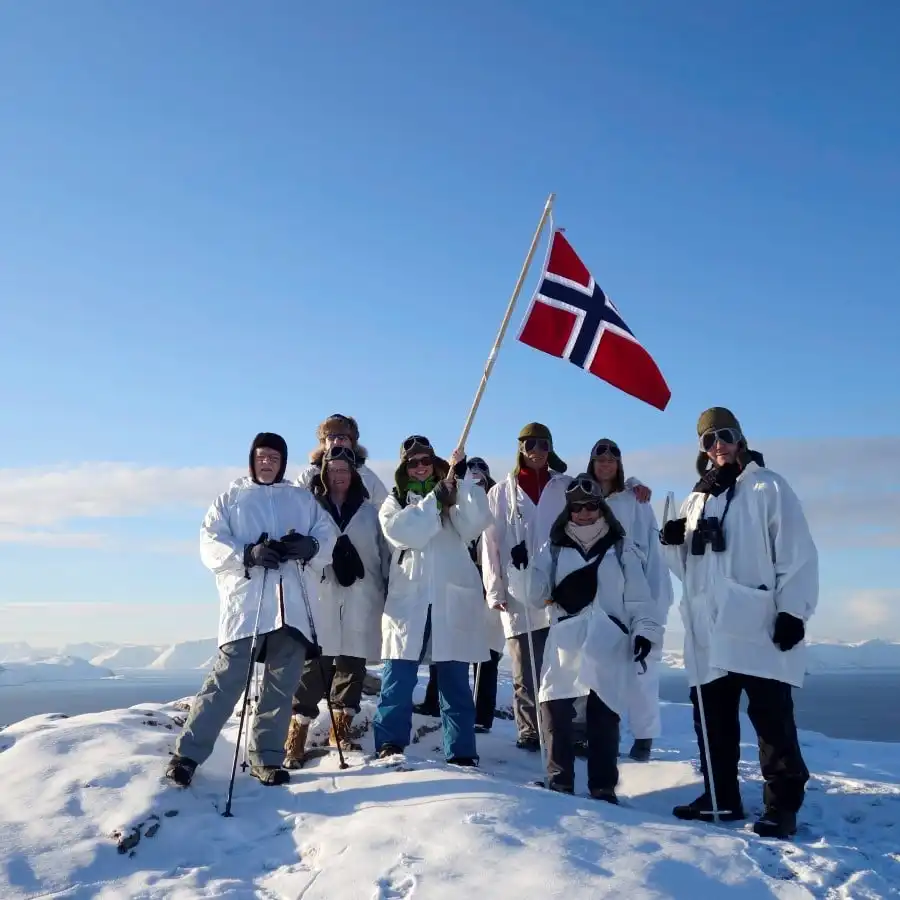 A group of 8 people in matching white jackets are waiving a Norwegian flag on top of a mountain top in Northern Norway