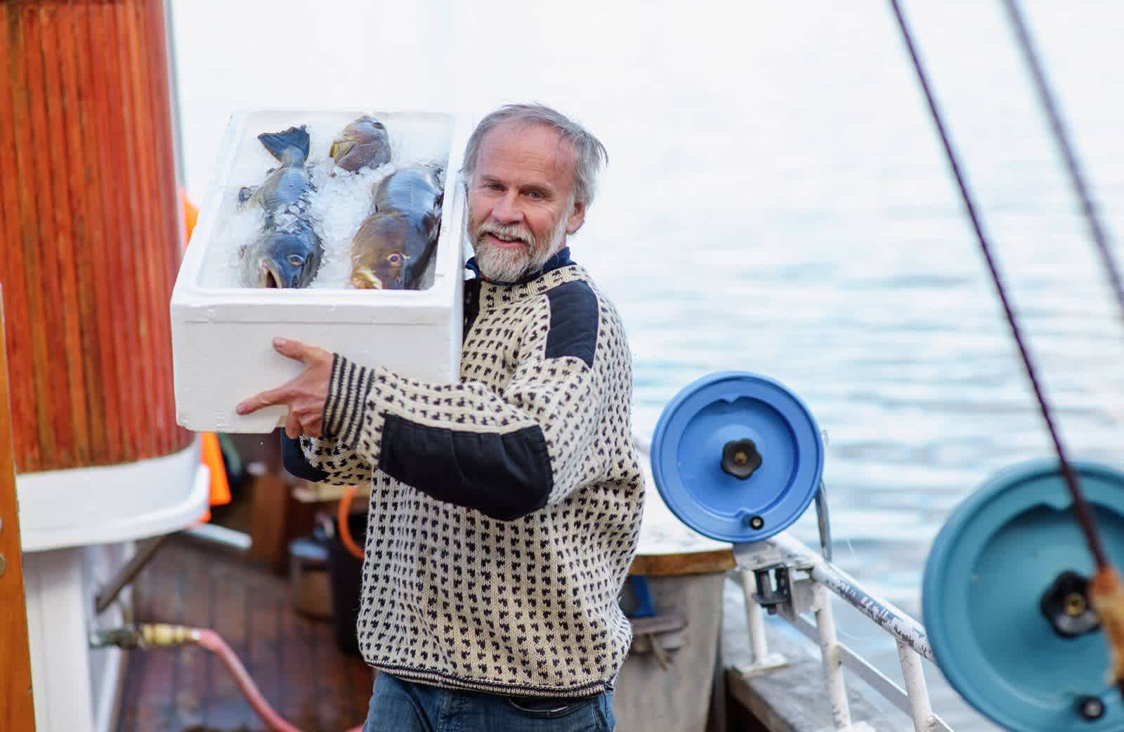 A fisherman holds a polystyrene box full of ice. At the top are three freshly caught cod, which are on their way to Hurtigruten's kitchen