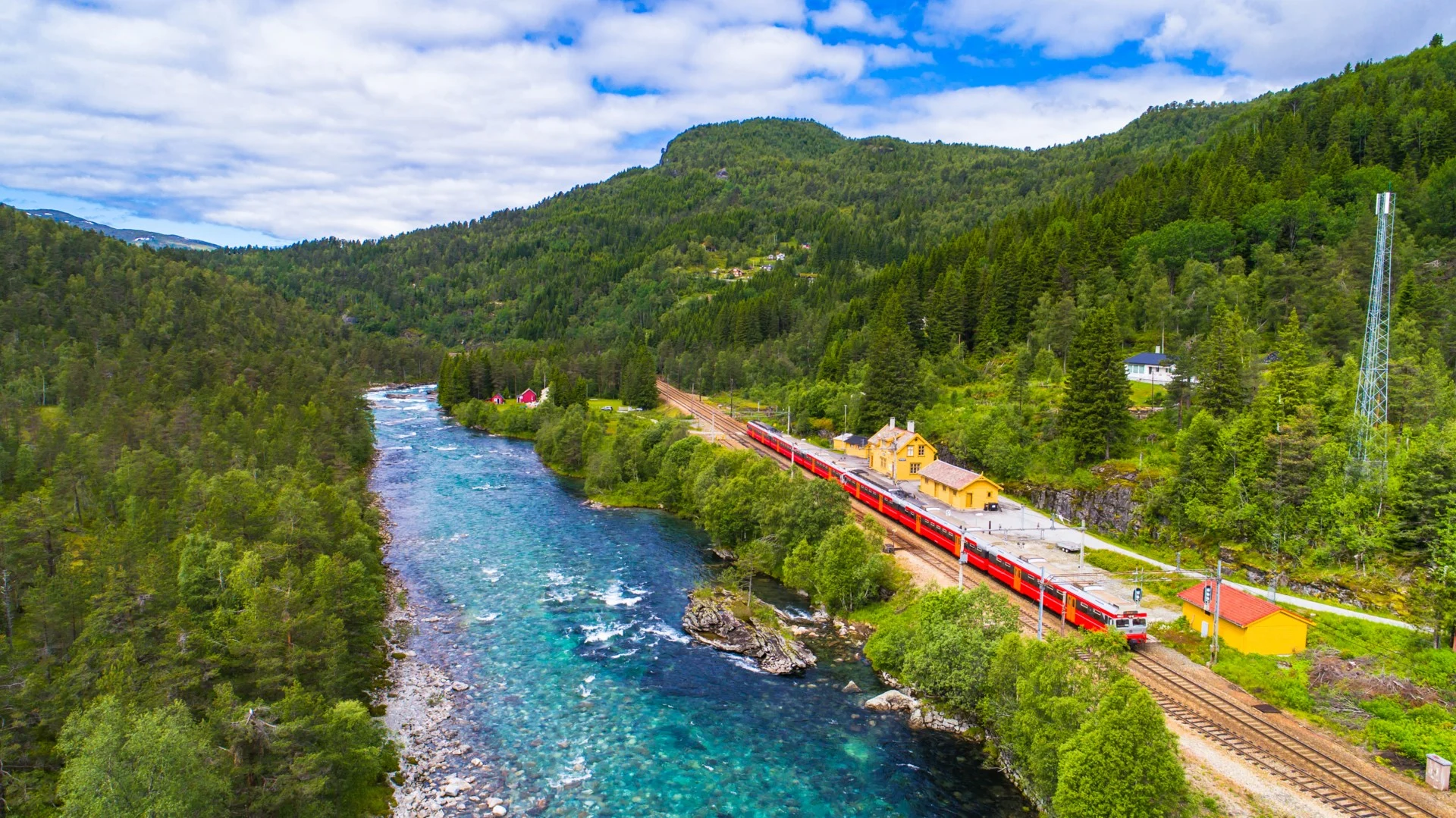 At a train station in Norway. The train has stopped at the station, and next to it flows a transparent blue river. It is summer season and the trees are green and fresh