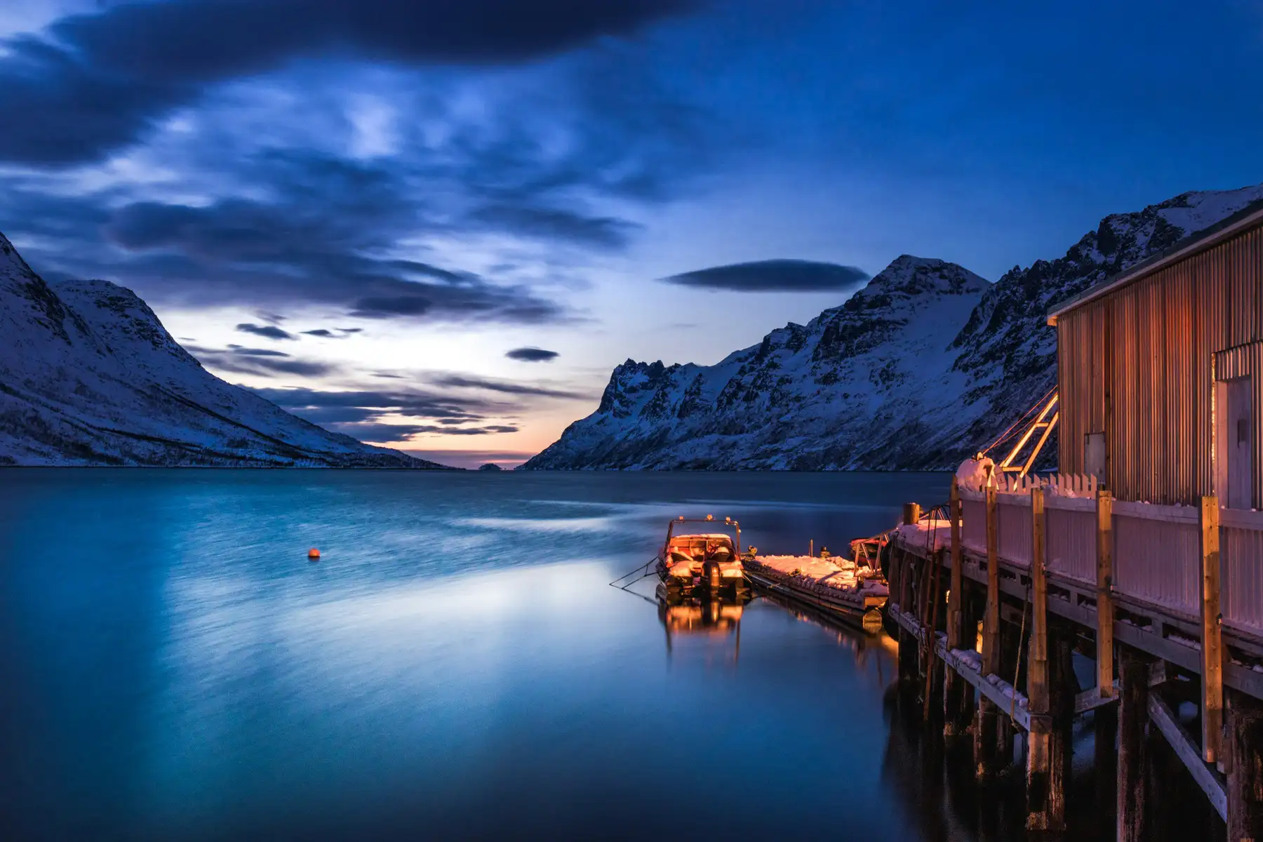 A traditional wooden boathouse glowing with warm amber light stands on stilts at the edge of a glassy fjord near Tromsø, Northern Norway, at winter twilight. A small snow-laden boat is moored at the adjacent jetty, its reflection shimmering in the still blue water. Snow-covered mountains rise steeply on both sides of the fjord, converging toward a faint orange glow on the horizon beneath a brooding, cloud-streaked sky.