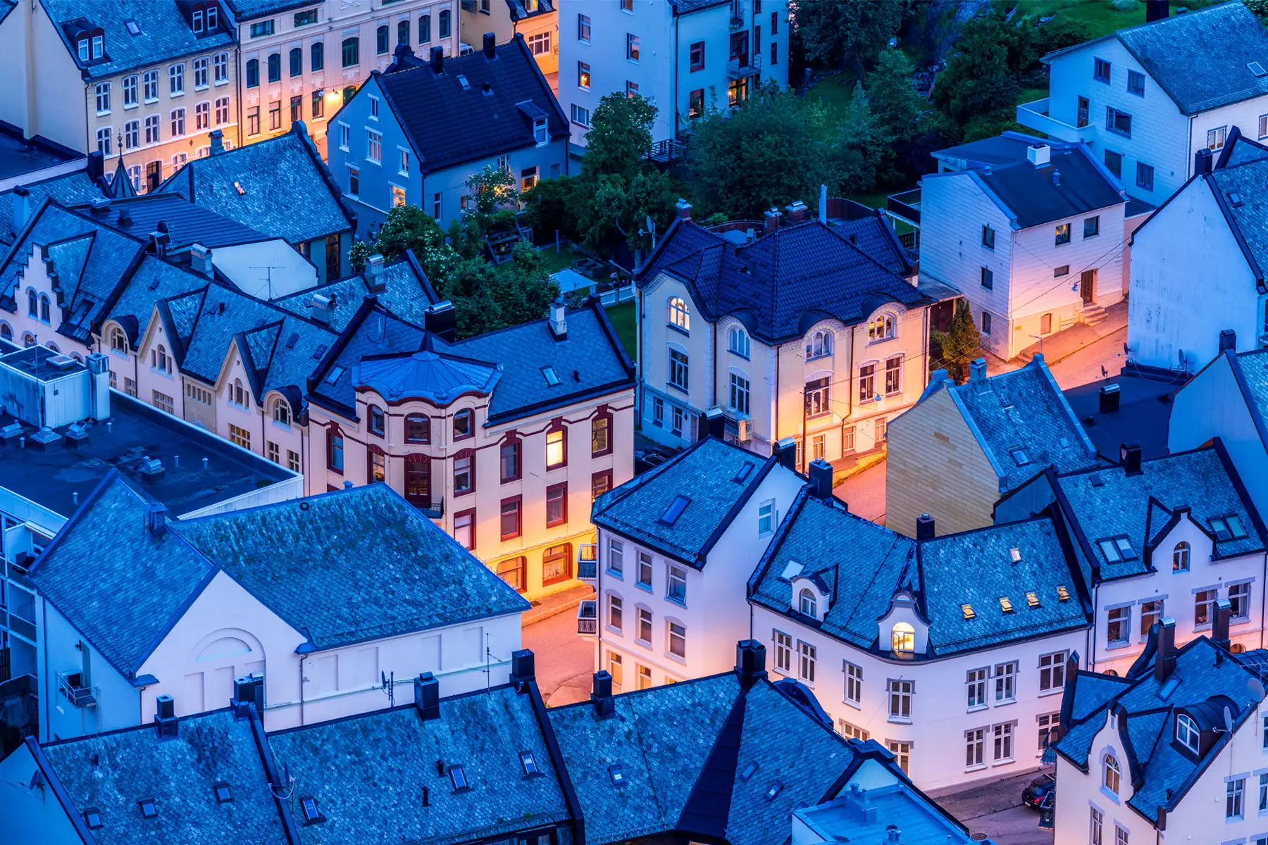 Bird's-eye view over the densely packed Art Nouveau buildings of Ålesund, Norway, photographed during blue hour. Slate-grey rooftops and white-rendered facades are bathed in cool twilight tones, while warm amber light spills from windows and narrow streets below.