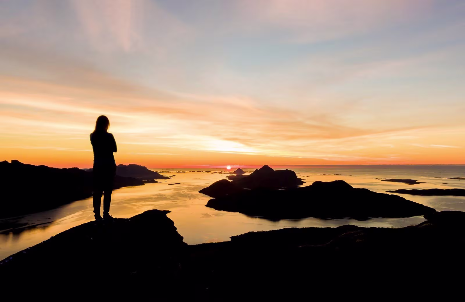 A young girl have hiked to the top of a peak, enjoying the view of the ocean, mountains and the midnight sun.