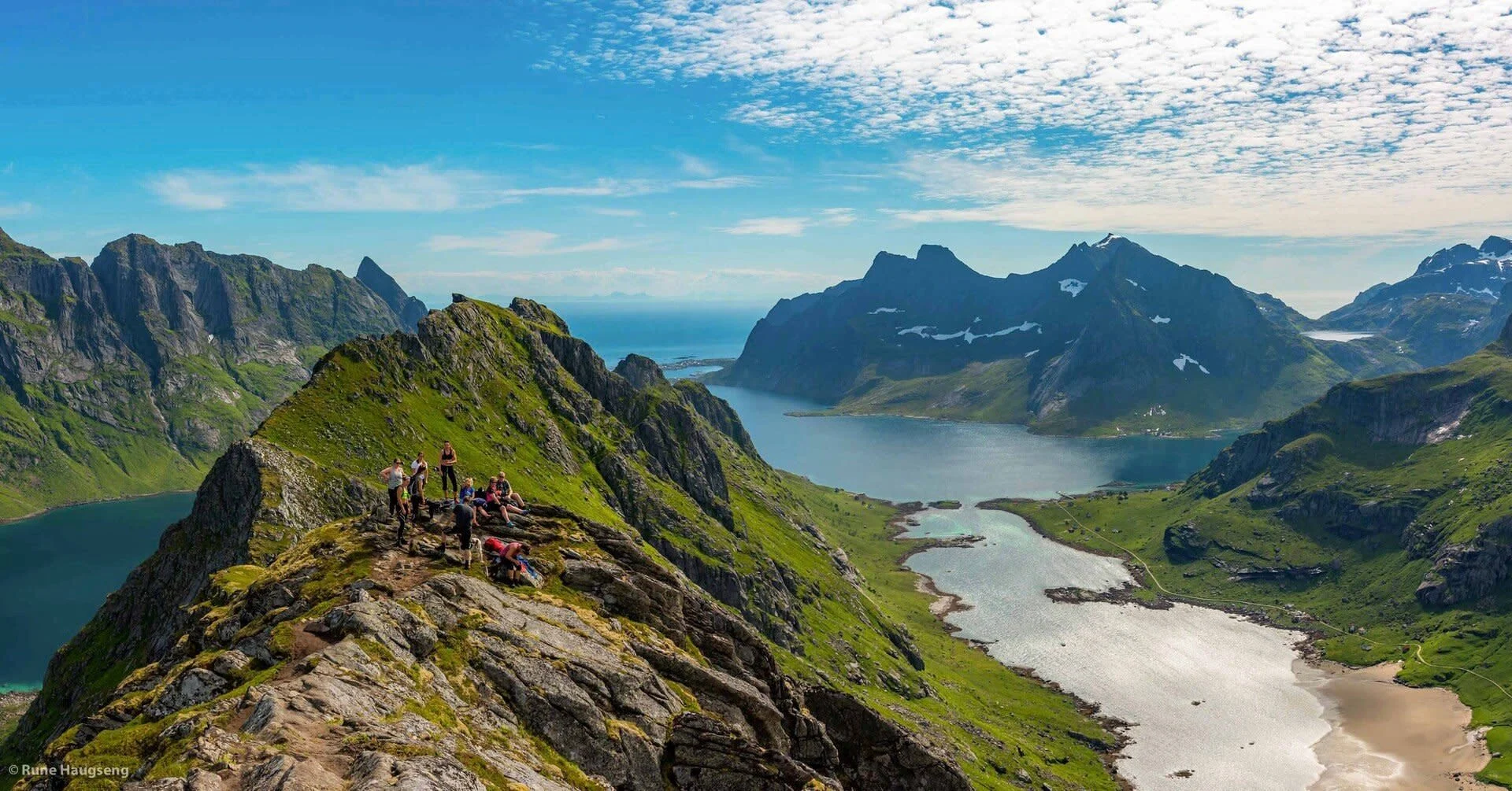Hikers on a mountain ridge overlooking a fjord with emerald waters and rugged peaks.