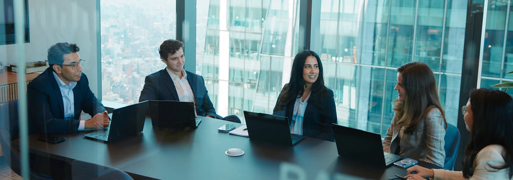Five people in a board room talking in front of a wall of windows