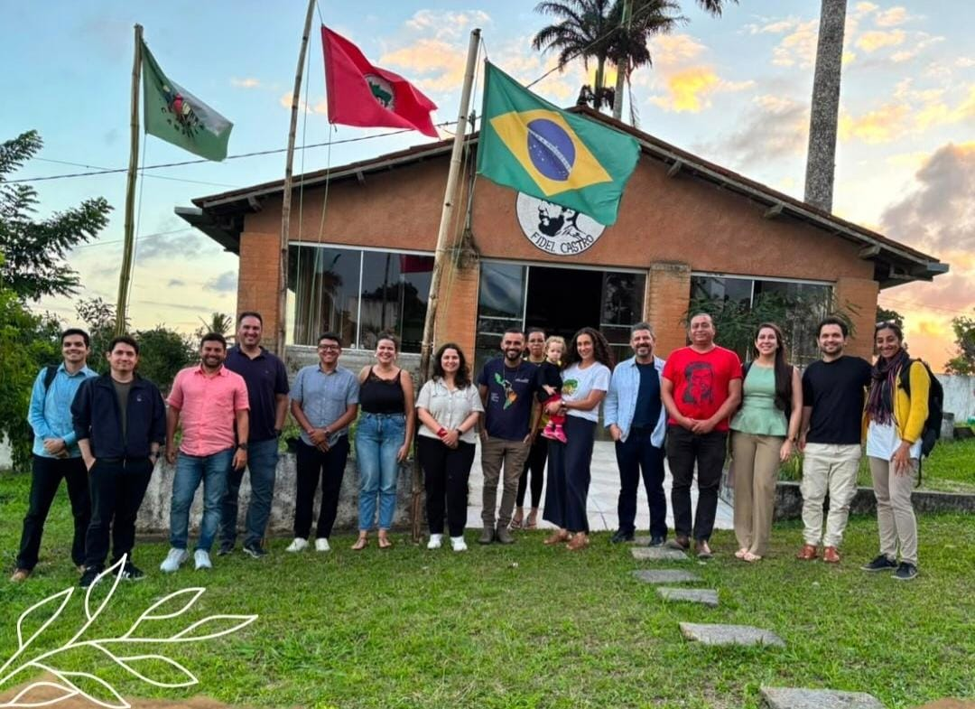 Fotografia de um grupo de pessoas reunido ao ar livre, em frente a uma edificação.  O grupo posa para a foto sobre uma área gramada, com árvores e vegetação ao redor, sob o céu ao entardecer.