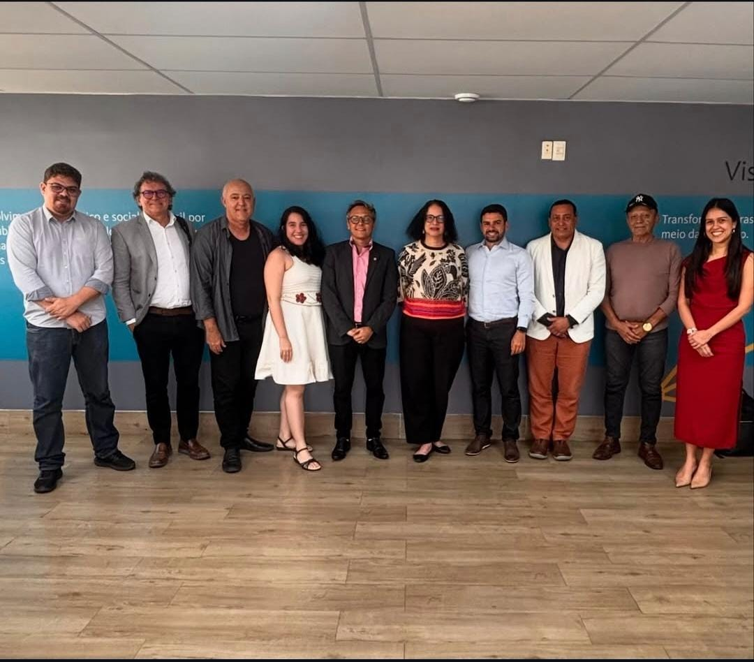 Grupo de dez pessoas em pé, lado a lado, posando para foto em ambiente interno. Ao fundo, parede cinza com faixa azul.