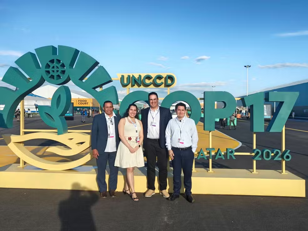 Quatro pessoas posam diante da instalação “UNCCD COP17 Ulaanbaatar 2026”, em área externa do evento. Ao fundo, aparecem pavilhões, estruturas da conferência e céu azul.